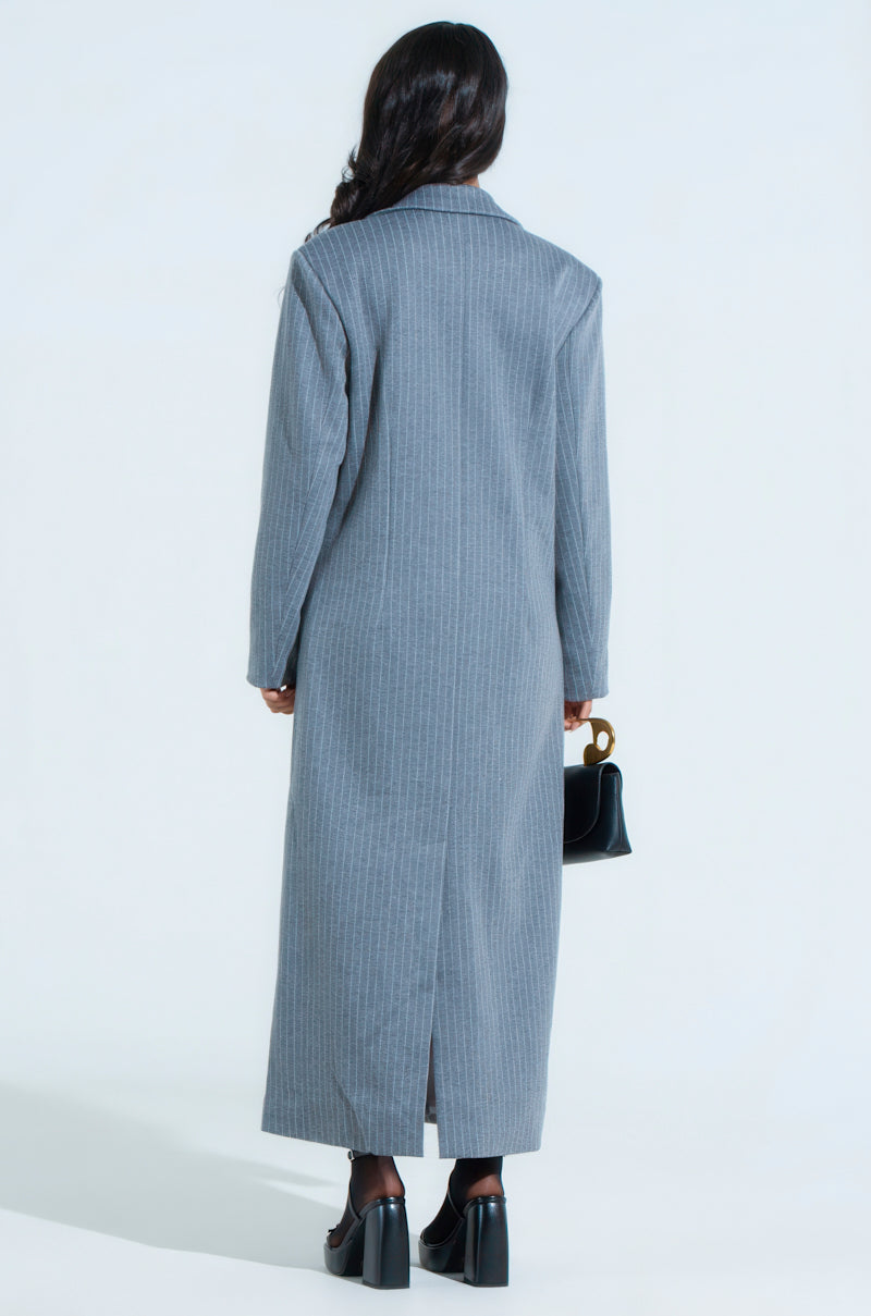 Rear view of a person with long hair wearing the "BUSINESS MEETS PLEASURE OVERSIZED TRENCH" in gray pinstripe, paired with black heeled shoes and holding a small black handbag. The coat features a back slit against a plain white background.