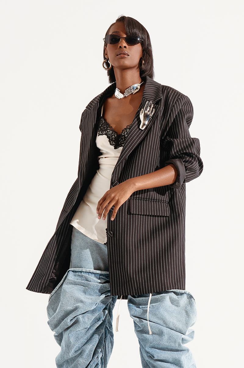 A person wearing oversized, baggy jeans, a white lace-trim camisole, and the ELLA OVERSIZED PINSTRIPE LONGLINE BLAZER adorned with a silver hand-shaped brooch. They accessorize with dark sunglasses and a chunky silver choker necklace, striking a confident pose against a plain background.