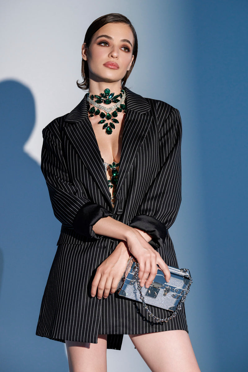 A woman in the ELLA OVERSIZED PINSTRIPE LONGLINE BLAZER, a green statement necklace, and a blue patterned handbag stands against a blue and white background, gazing slightly to the side.
