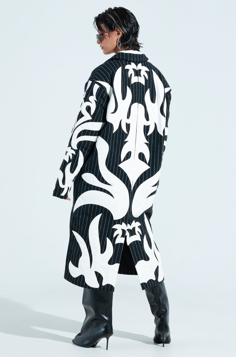 A person wearing the PEPPER PINSTRIPE TRENCH, featuring oversized longline style with bold white abstract patterns, stands back to the camera. They have short hair, sunglasses, and black knee-high boots against a plain, light backdrop that enhances their unique style.