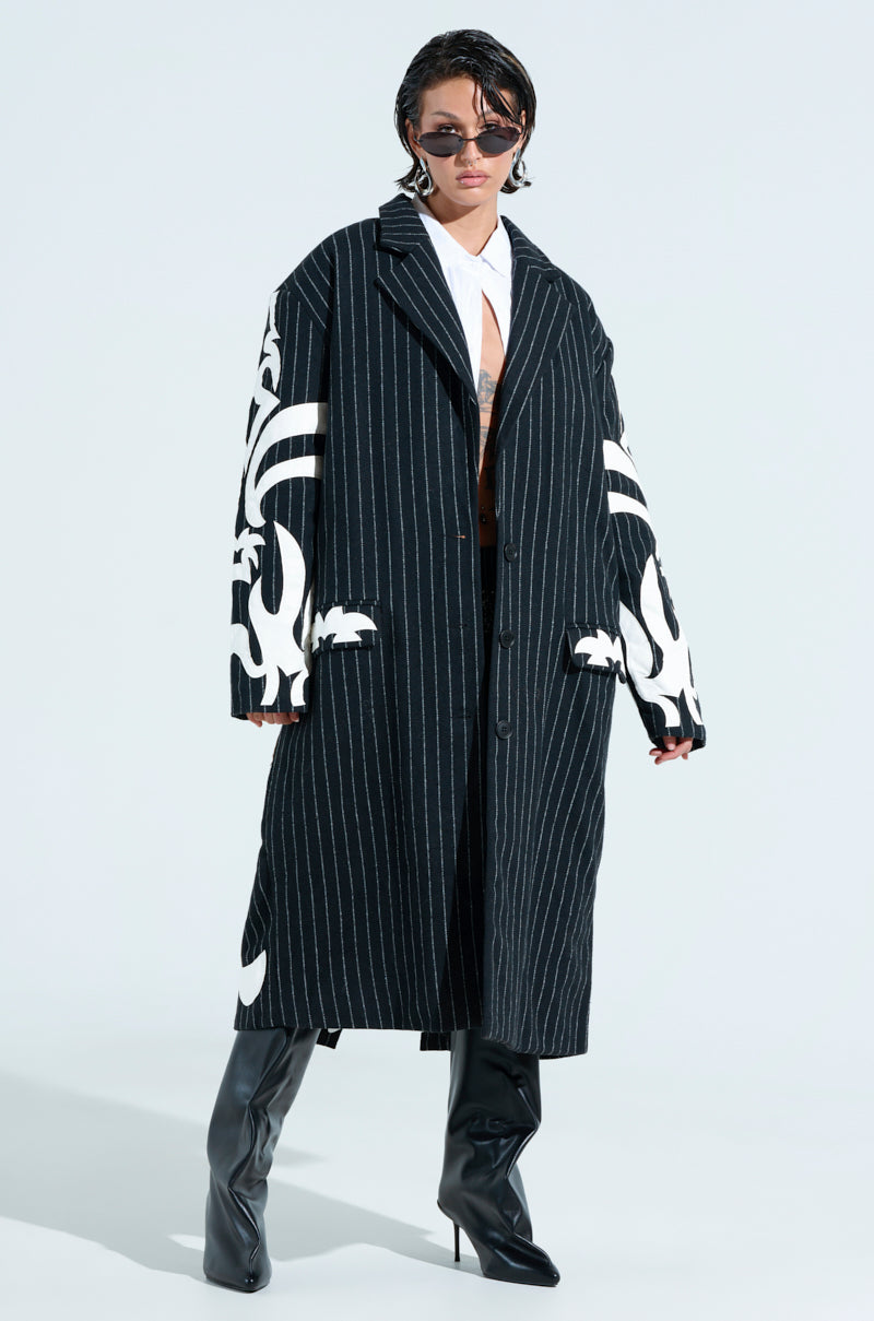 A person wears the PEPPER PINSTRIPE TRENCH with white patterns over a classic white shirt and tie, accessorized with sunglasses and black knee-high boots, making a striking impression against a plain, light background.