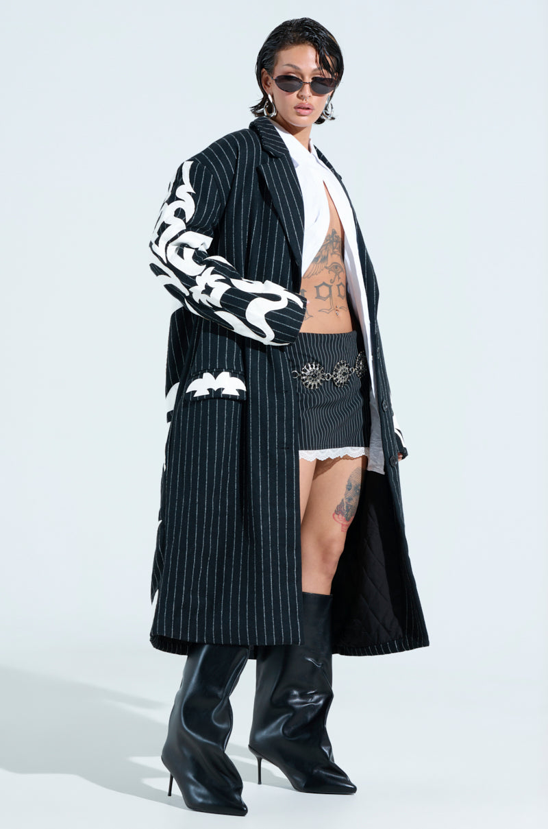 Against a plain background, a person wears sunglasses and the Pepper Pinstripe Trench with bold white patterns, complemented by a white shirt, black shorts, high-heeled boots, and tattoos on their chest and legs.