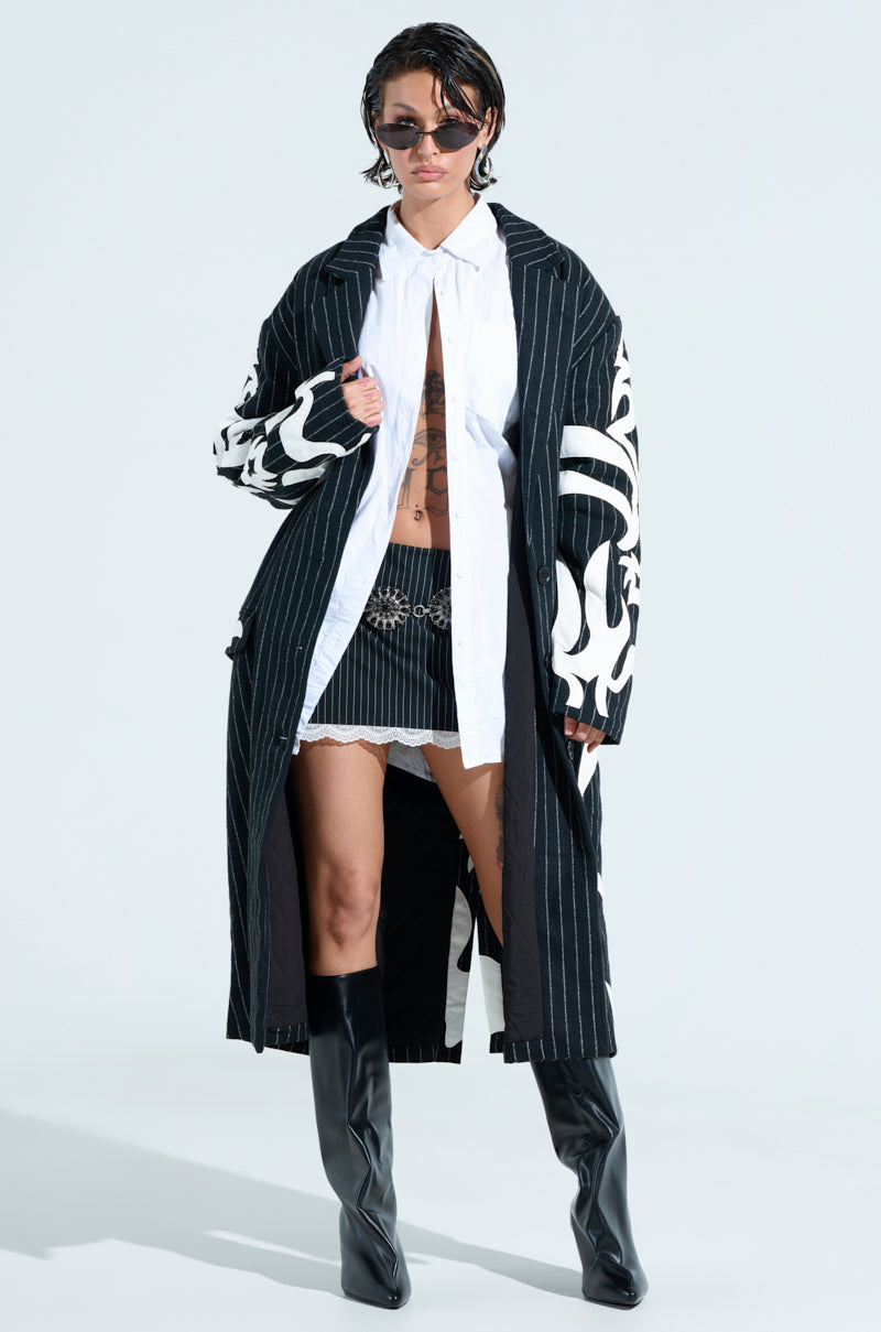 A person dons the PEPPER PINSTRIPE TRENCH with an oversized fit, white patterns, a white shirt, and a black skirt. They accessorize with sunglasses and black knee-high boots, revealing a brown tattoo on their chest. The plain backdrop complements the embroidered faux leather patches enhancing their style.