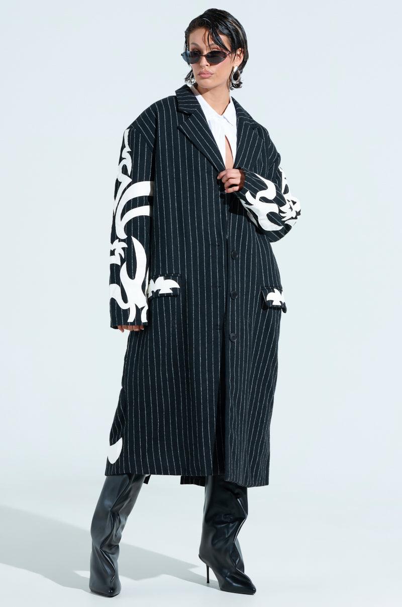 Wearing a black PEPPER PINSTIPE TRENCH with striking white patterns, short hair, sunglasses, and black heeled boots paired with a white top, the person confidently embodies modern style.