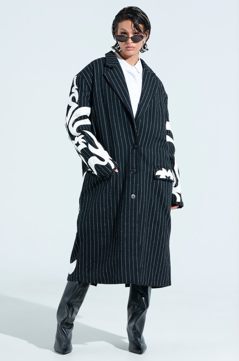 Wearing an oversized PEPPER PINSTIPE TRENCH with embroidered faux leather sleeve patches, a short-haired person poses confidently in sunglasses, knee-high black boots, against a plain, light background.