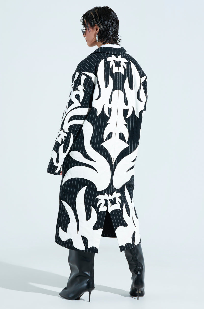 A person confidently wears the oversized PEPPER PINSTRIPE TRENCH with bold black and white designs. High black boots and sunglasses complete the look, while a light gray background highlights the striking ensemble.