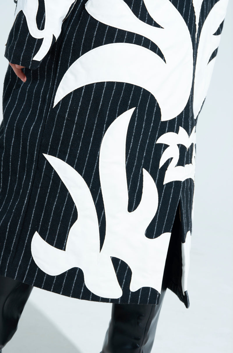 The image features a person wearing the PEPPER PINSTRIPE TRENCH, an oversized longline coat with black pinstripes and bold white abstract plant designs, paired with black leather pants. Photographed from the side to showcase the entire ensemble.