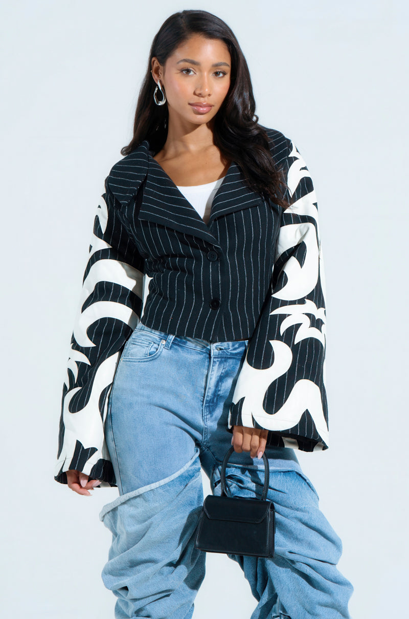 A person poses in a stylish outfit featuring the HEART ON FIRE EMBROIDERED BLAZER with flared sleeves, a white top, and light blue layered jeans. They hold a small black handbag and wear hoop earrings.