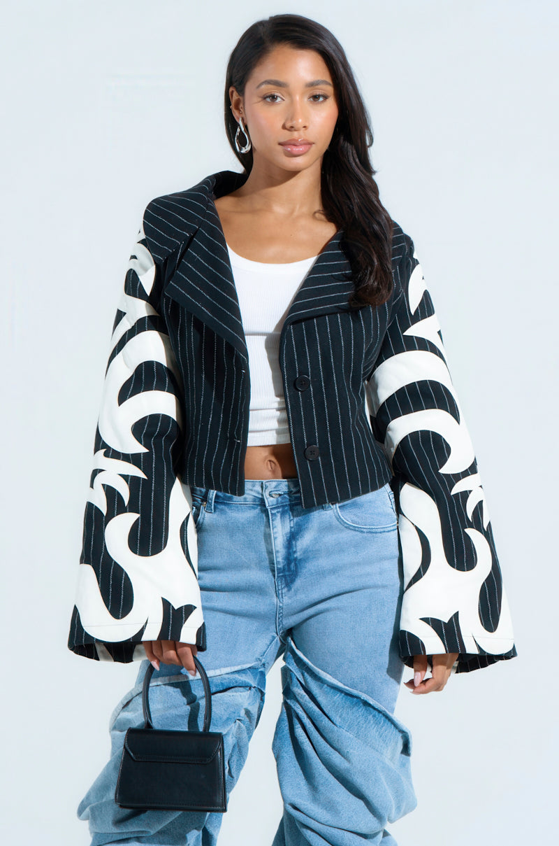 A person with long dark hair sports the HEART ON FIRE EMBROIDERED BLAZER, with abstract sleeves over a white top. They pair this ensemble with blue jeans and hold a small black handbag.