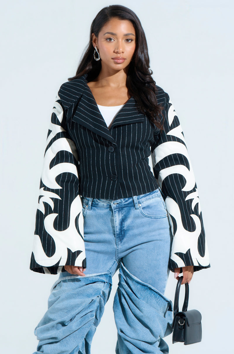 A woman with long hair wears a HEART ON FIRE EMBROIDERED BLAZER, paired with a crisp white top and baggy blue jeans. She carries a small black handbag and sports hoop earrings, standing against a light background.