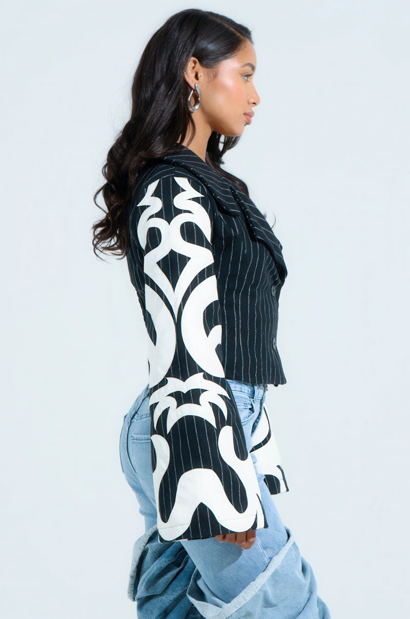 A woman with long dark hair is in profile against a light background, wearing the HEART ON FIRE EMBROIDERED BLAZER with flared sleeves and light blue jeans. Hoop earrings highlight her serious expression, enhancing her striking ensemble.