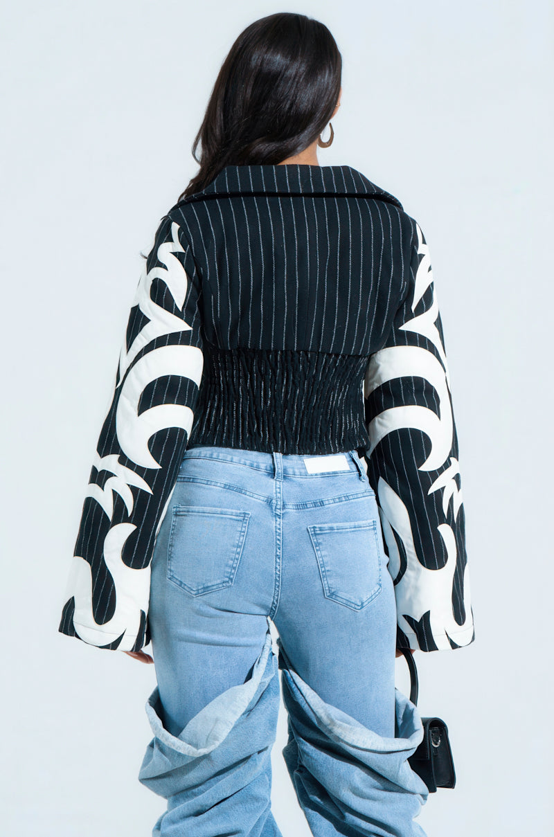 Facing away, a person with long dark hair dons the HEART ON FIRE EMBROIDERED BLAZER, pairing it with light blue jeans featuring a unique layered design. They hold a small black handbag against the light gray background.
