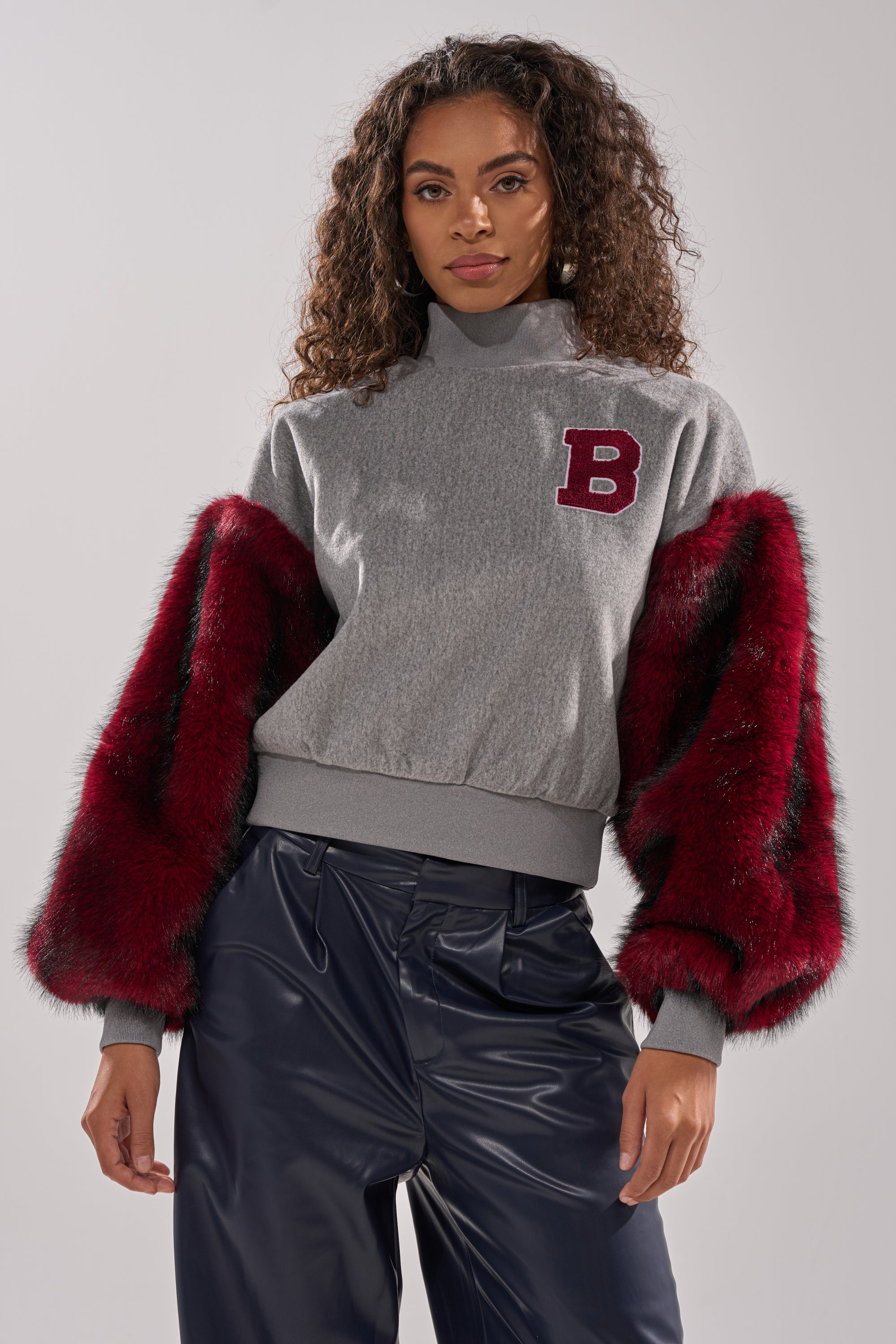 A woman models the NEW EAST SIDE FAUX FUR SLEEVE SWEATSHIRT with gray faux fur sleeves and a red "B" patch, hands in pockets, paired with shiny dark blue pants—an oversized streetwear look against a light gray background.