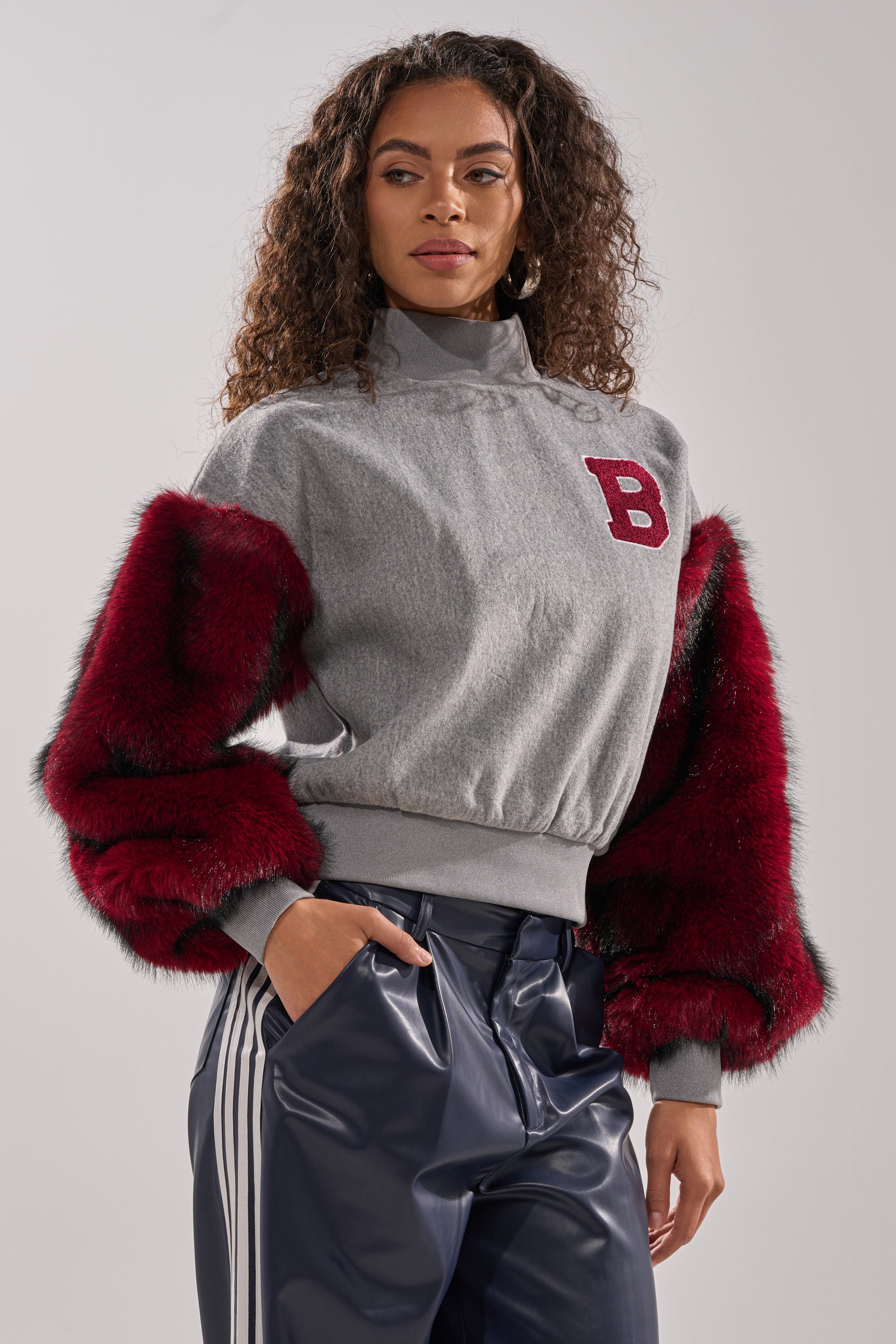 A woman with curly hair models the NEW EAST SIDE FAUX FUR SLEEVE SWEATSHIRT, showcasing its red "B" patch and plush red sleeves, paired with shiny navy blue track pants for a standout streetwear look against a plain background.