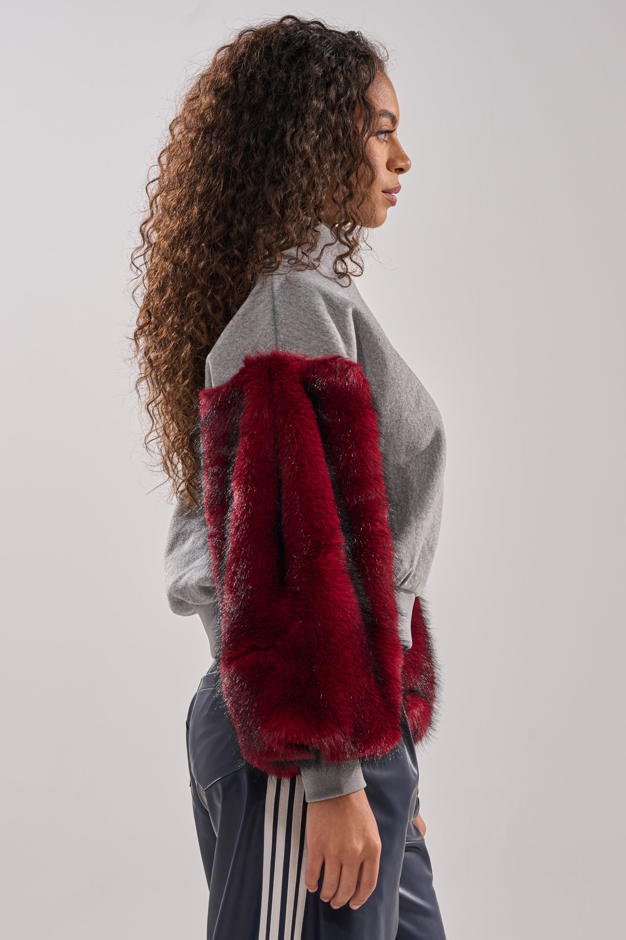 A woman with long curly hair models the NEW EAST SIDE FAUX FUR SLEEVE SWEATSHIRT—an oversized grey pullover with red faux fur sleeves and hem—paired with dark pants with white side stripes, standing in profile against a light grey background.