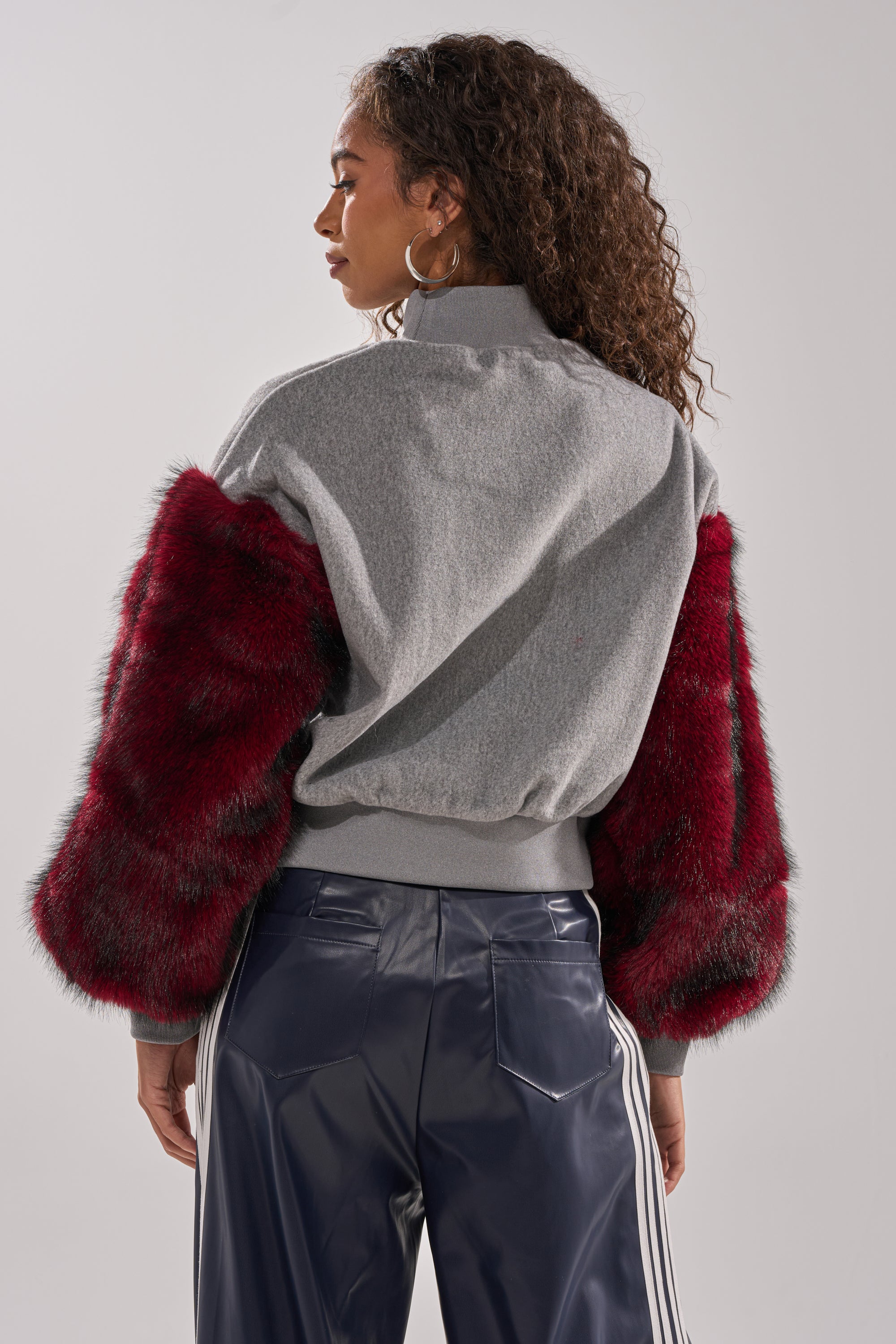 A woman with curly hair, seen from behind, models streetwear style in the NEW EAST SIDE FAUX FUR SLEEVE SWEATSHIRT paired with navy pants featuring white side stripes against a plain background.