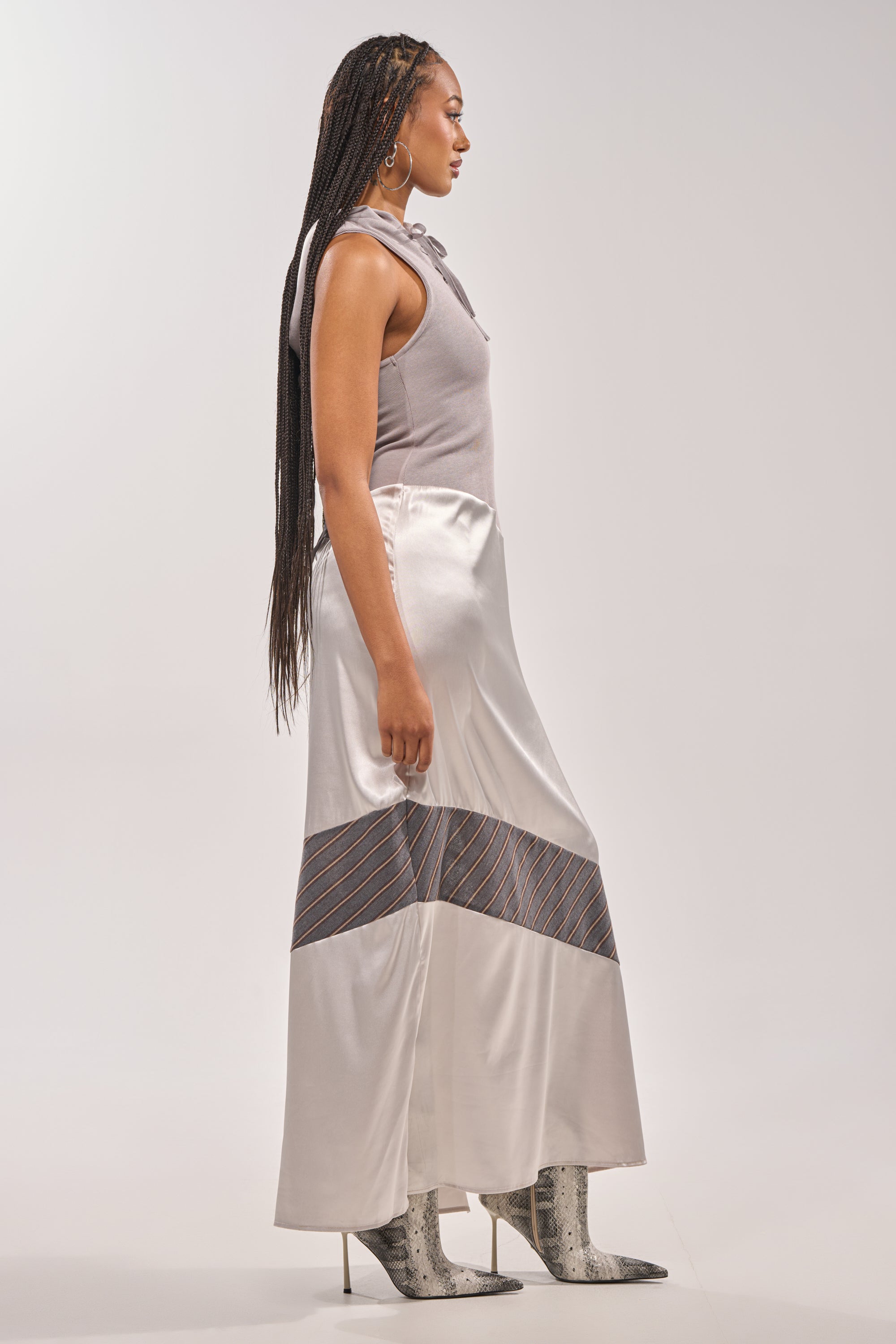 A woman with long braids stands in profile, wearing the BONDI PATCHWORK HOODED MAXI DRESS IN GREY against a plain light background.