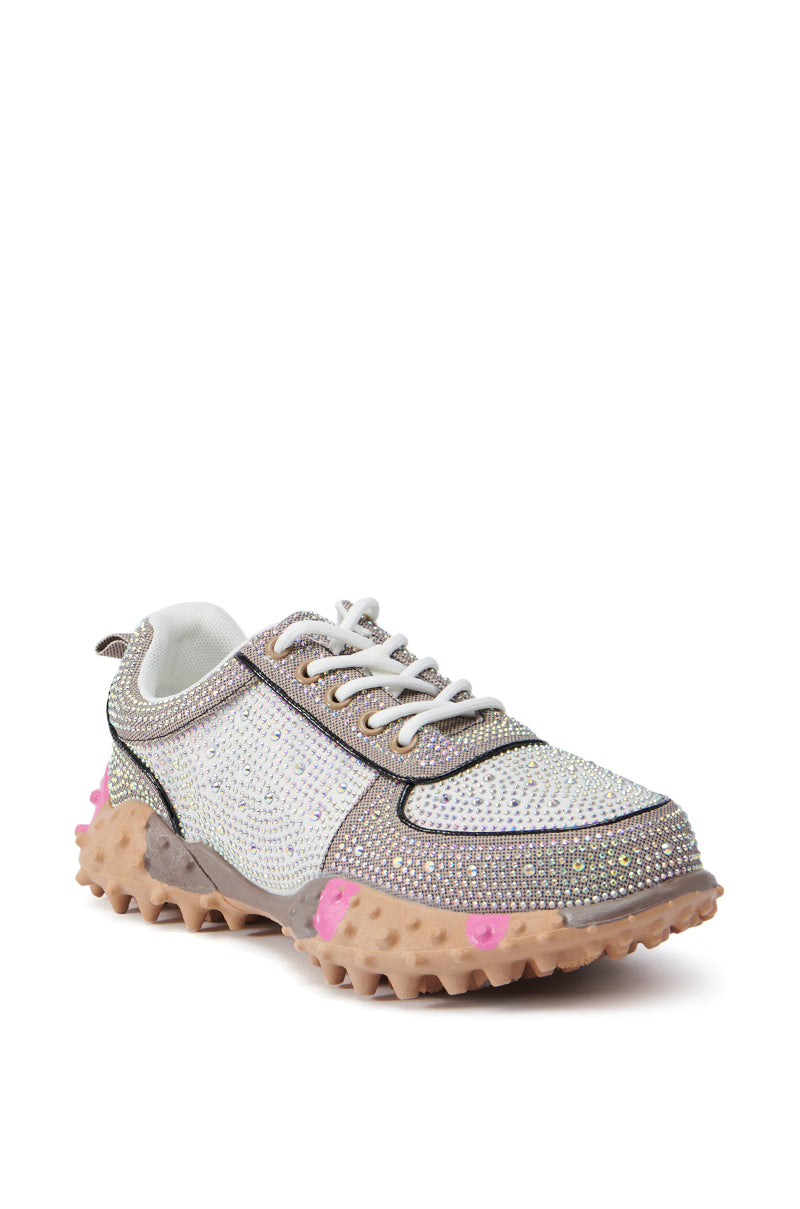 The AZALEA WANG ISABEAU BROWN MULTI SNEAKER is a rhinestone-embellished white sneaker that features grey accents, white laces, and a thick, rugged beige sole. The distinct textured sole has a knobby surface with hints of pink detailing. This sporty yet flashy design is further enhanced by its subtle color block pattern.