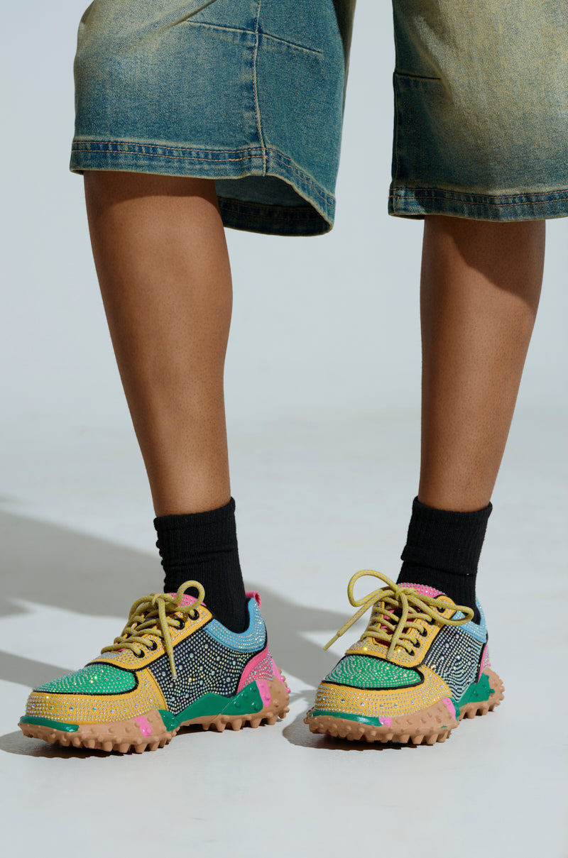 Standing on a neutral background, an individual sports AZALEA WANG ISABEAU EMBELLISHED MULTI SNEAKERS with rhinestones and green and pink accents. Paired with black socks and cropped denim shorts, the cleated rubber sole adds a stylish touch.