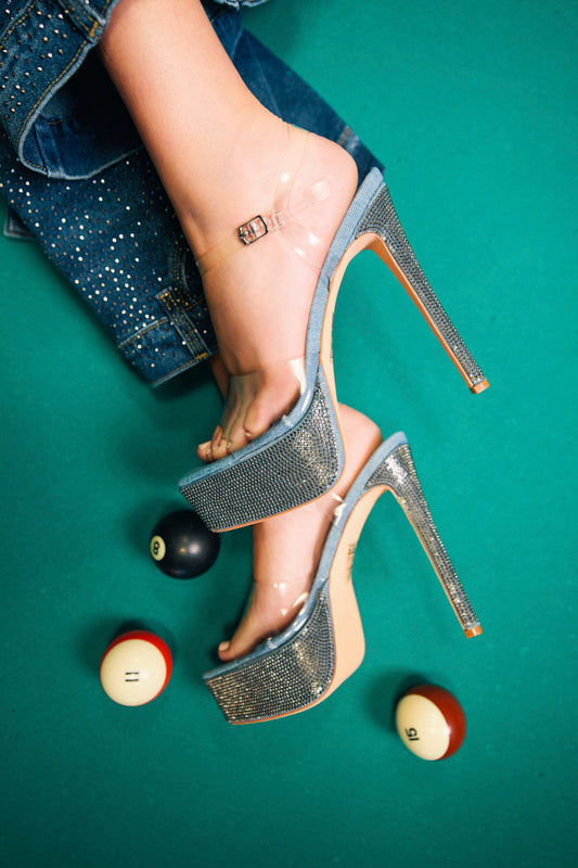 Someone wears the AZALEA WANG ISAURA DENIM PLATFORM RHINESTONE SANDAL, resting their sparkly rhinestone heels with clear straps on a green pool table with billiard balls (8, 11, and 15). Rhinestone-accented denim pants are partially in view.