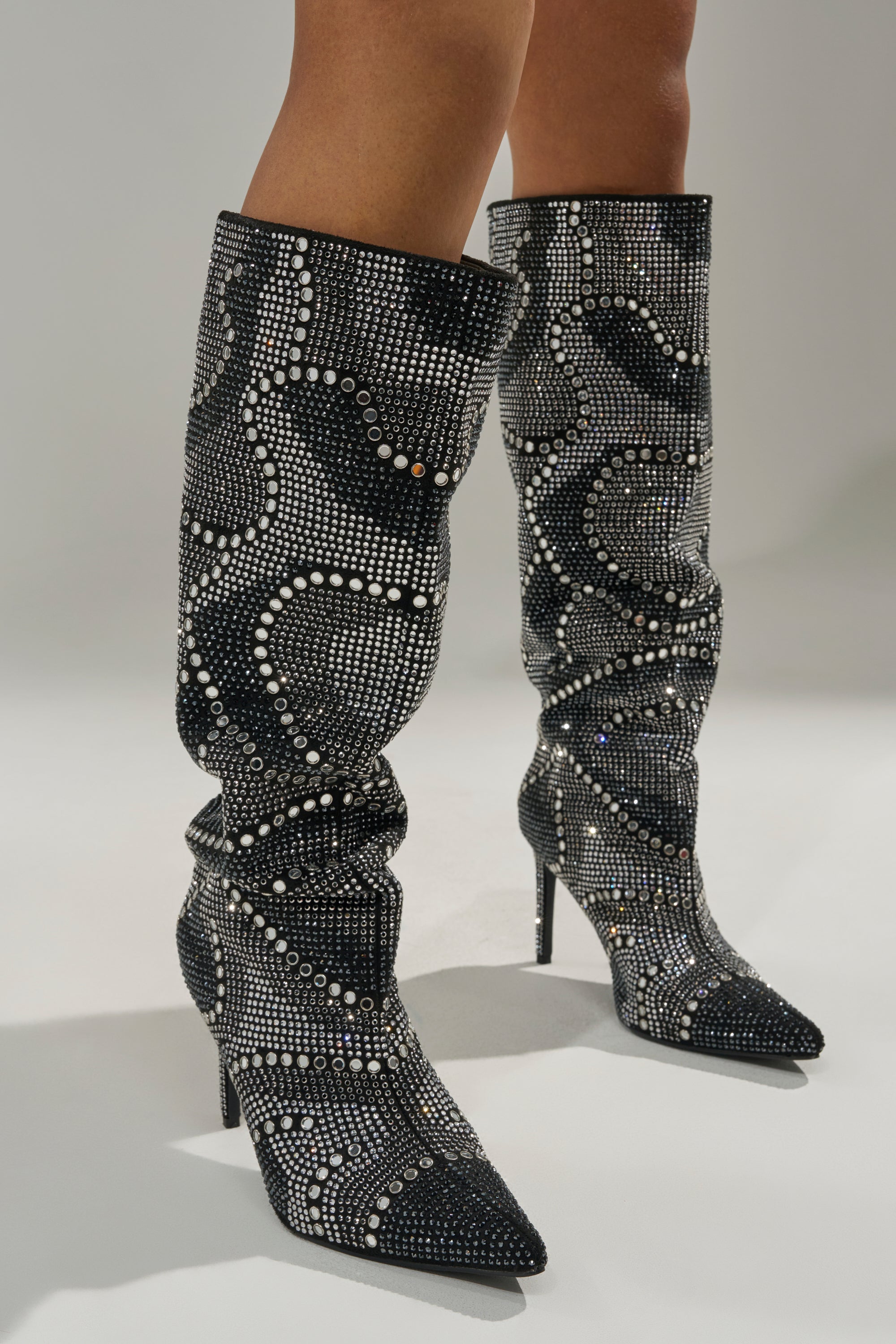 A person wearing AZALEA WANG ISILDA BLACK MULTI SEQUIN STILETTO BOOTS, featuring knee-high shimmering patterns, stands confidently on a light gray surface.