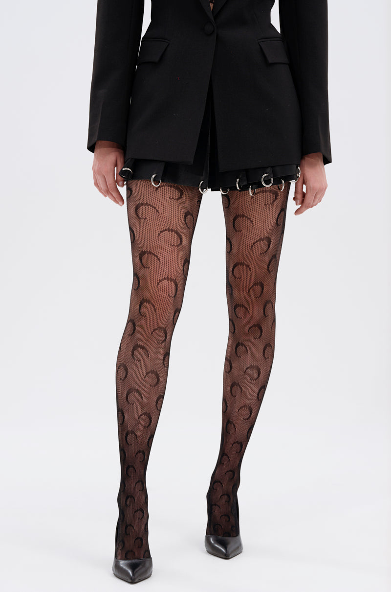 A person wearing crescent moon pattern AFTER MIDNIGHT BLACK TIGHTS, a black blazer, black mini skirt with metal rings, and black pointed-toe heels stands against a plain light background.