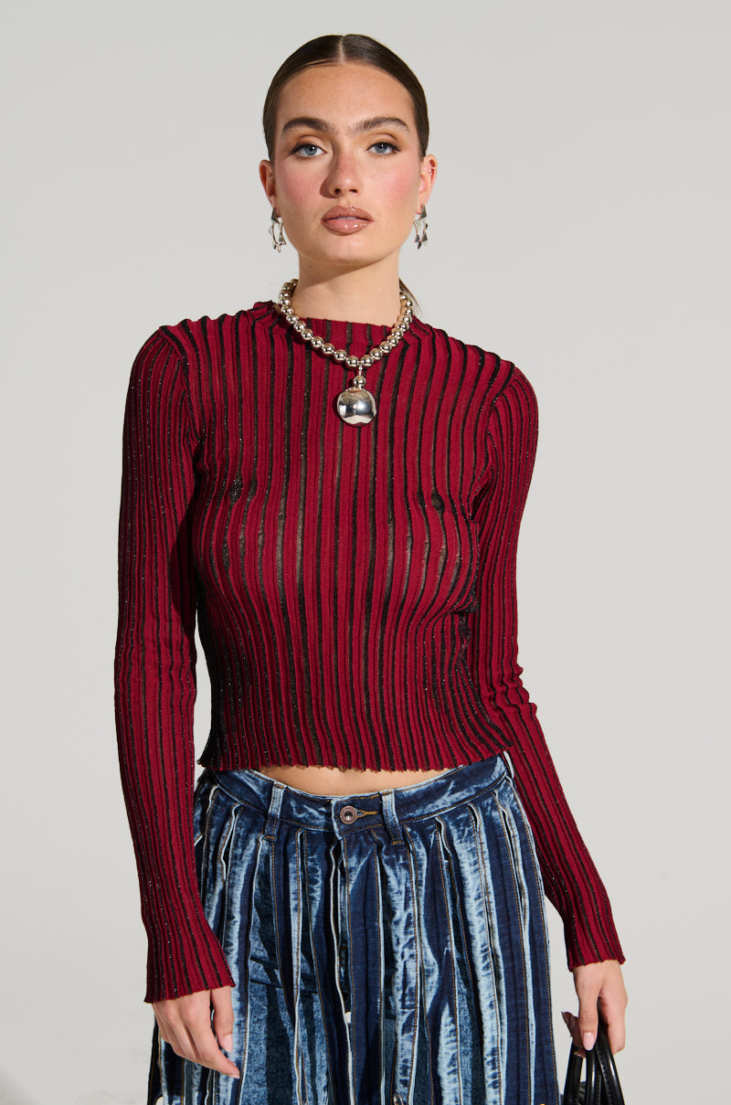 A person stands against a plain background, wearing the LOOKING SNATCHED STRIPED KNIT TOP IN RED paired with high-waisted blue velvet pants. Their hair is pulled back, complemented by earrings and a chunky chain necklace featuring a silver pendant.