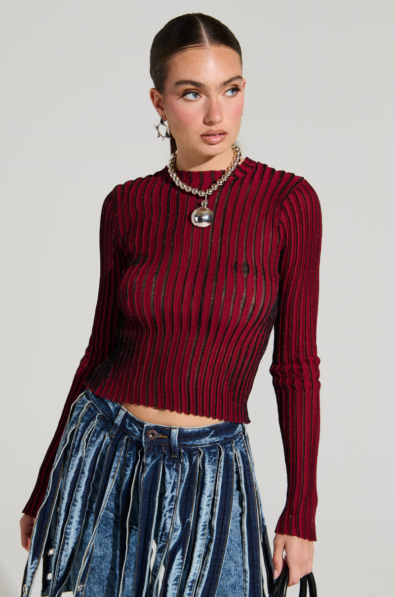 A woman with a sleek ponytail is wearing the LOOKING SNATCHED STRIPED KNIT TOP IN RED, which is fitted and long-sleeved. She pairs it with a denim skirt featuring dark and light blue vertical stripes, accessorizes with a pendant necklace, and holds a black handbag. She gazes to her right with a neutral expression.