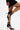 A person in a black mini dress stands on one leg, showing off the AZALEA WANG ITHYKA BLACK PATENT STILETTO PUMP with pointed toes and multiple buckle ankle straps against a plain white background.