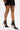A person stands on a white background wearing a black skirt and the AZALEA WANG ITHYKA BLACK PATENT STILETTO PUMP, featuring ankle straps and buckle details.