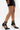 A person in a short black skirt and AZALEA WANG ITHYKA BLACK PATENT STILETTO PUMP heels with buckles stands against a plain white background.