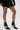 A person stands against a plain white background wearing a black mini skirt and AZALEA WANG ITHYKA BLACK PATENT STILETTO PUMP high-heeled shoes featuring multiple straps and a stiletto heel.