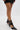 Wearing a short black skirt and sheer tights, a person stands on a white floor, highlighting the AZALEA WANG ITHYKA BLACK PATENT STILETTO PUMP with pointed toes, strap details, and glossy faux patent leather.