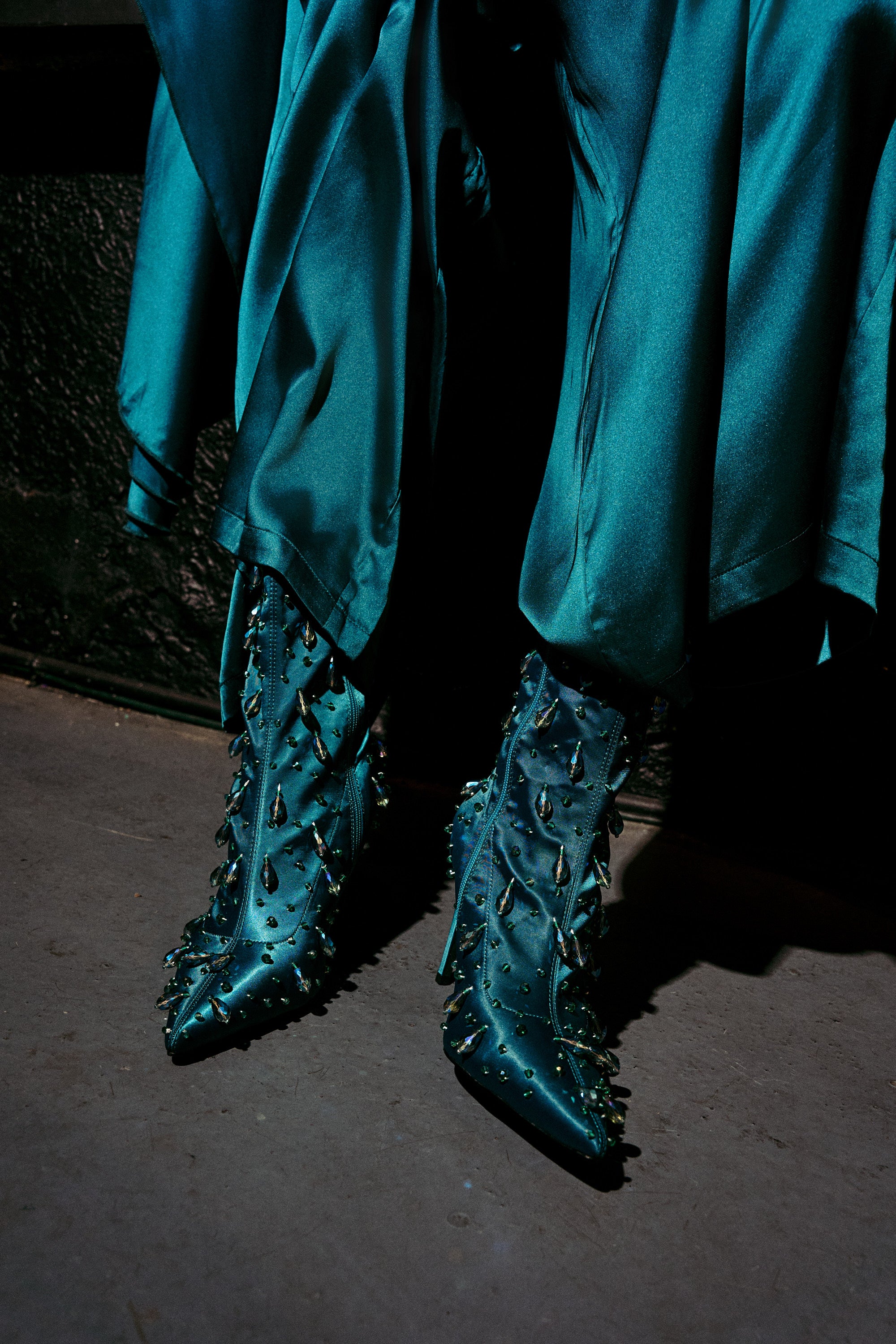 A person in a shiny teal dress stands on a concrete floor, showcasing the AZALEA WANG IZIDOR TEAL EMBELLISHED RHINESTONE BOOTIE with matching teal fabric and embellished rhinestone details.
