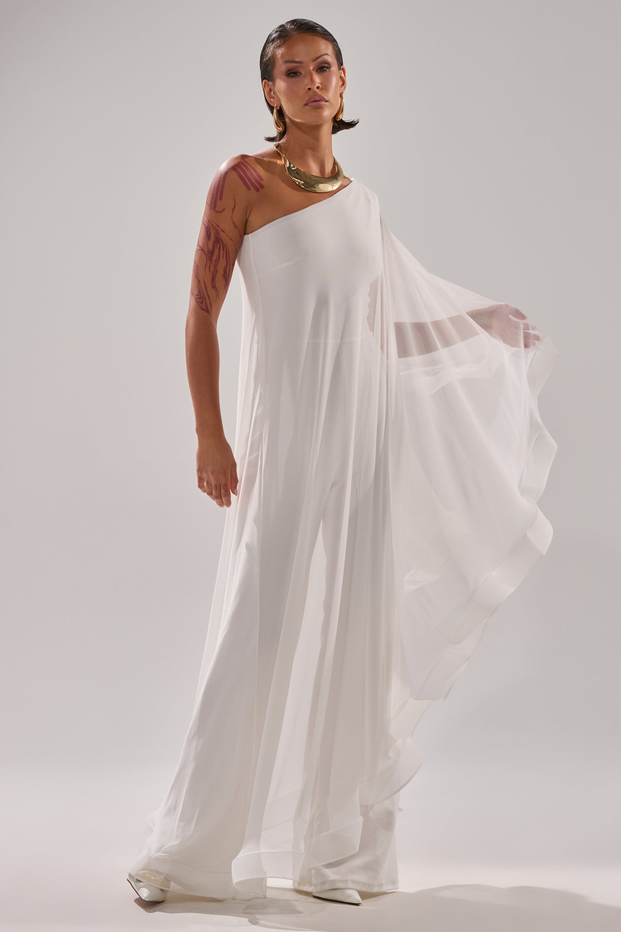 A person poses confidently in the SANDO MESH JUMPSUIT IN WHITE, featuring a flowing one-shoulder design. With slicked-back hair, gold jewelry, and visible red body art on one arm, they stand against a plain light background. 0