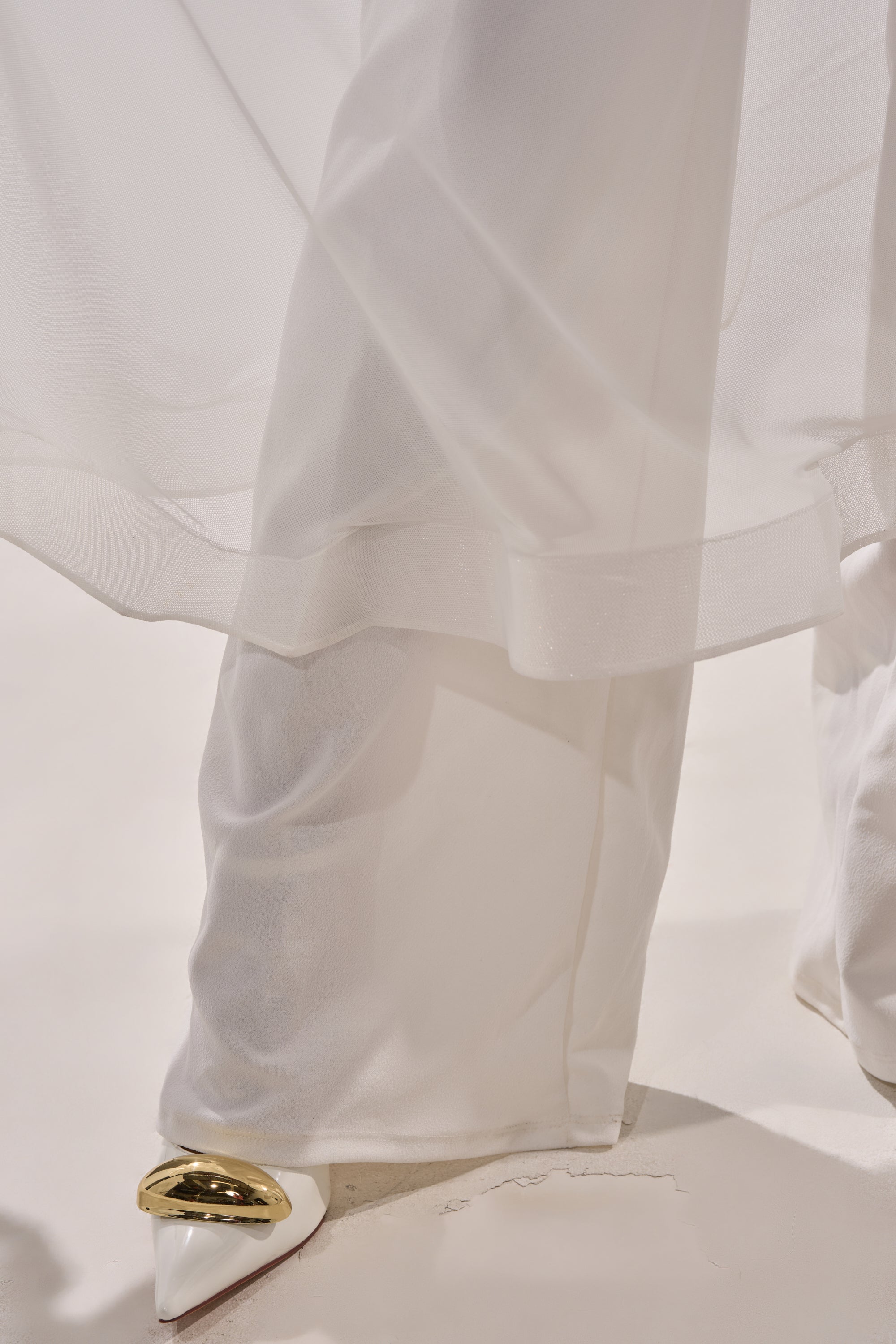 A close-up of the SANDO MESH JUMPSUIT IN WHITE shows sheer white fabric draping over legs, partially obscuring white pointed-toe shoes with gold accents, set against a light background. 9
