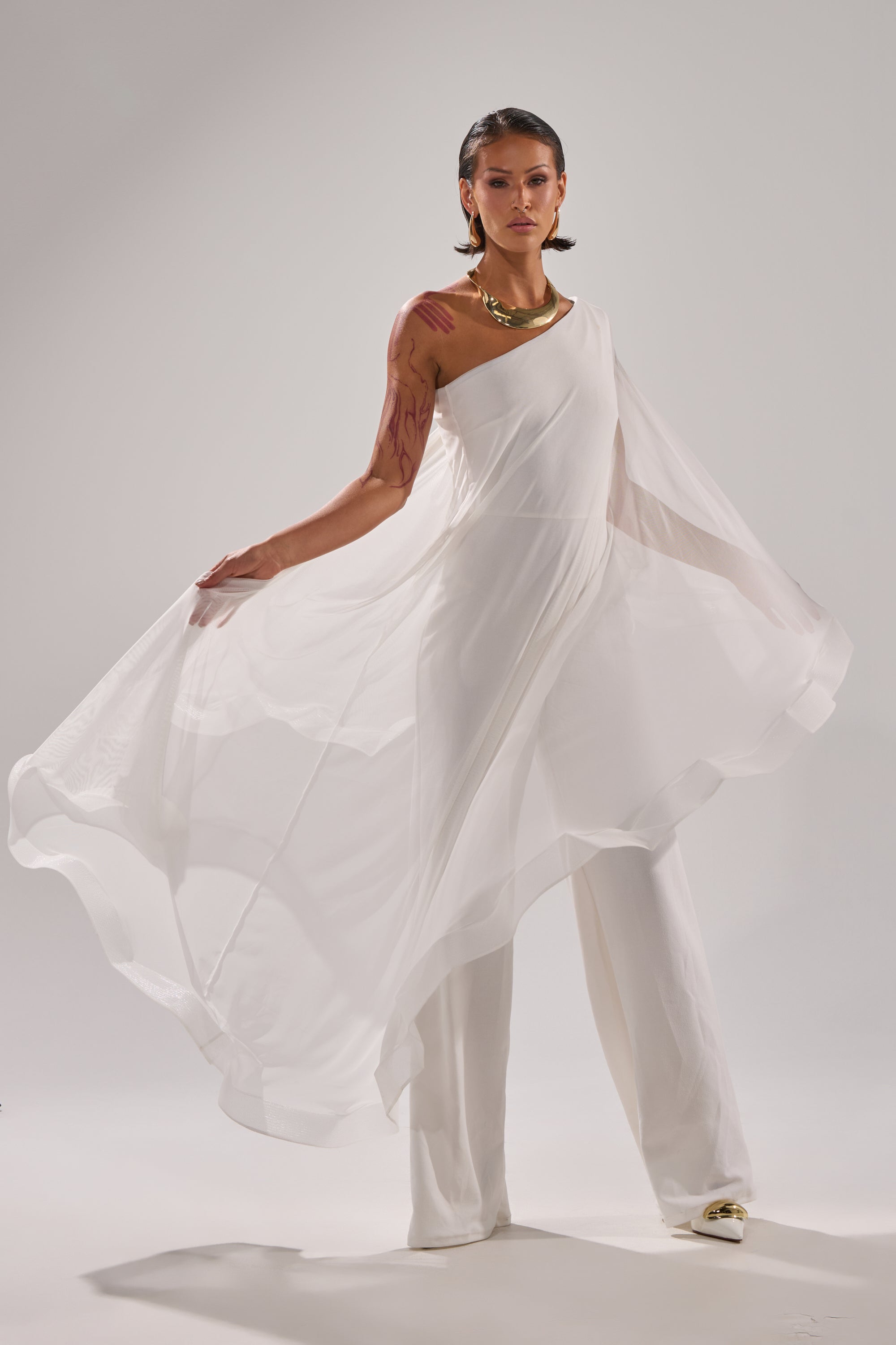 A woman models the SANDO MESH JUMPSUIT IN WHITE, featuring flowing mesh panels and white pants. She accessorizes with a gold necklace, her short hair styled simply, while her pose showcases the airy movement against a plain backdrop. 1