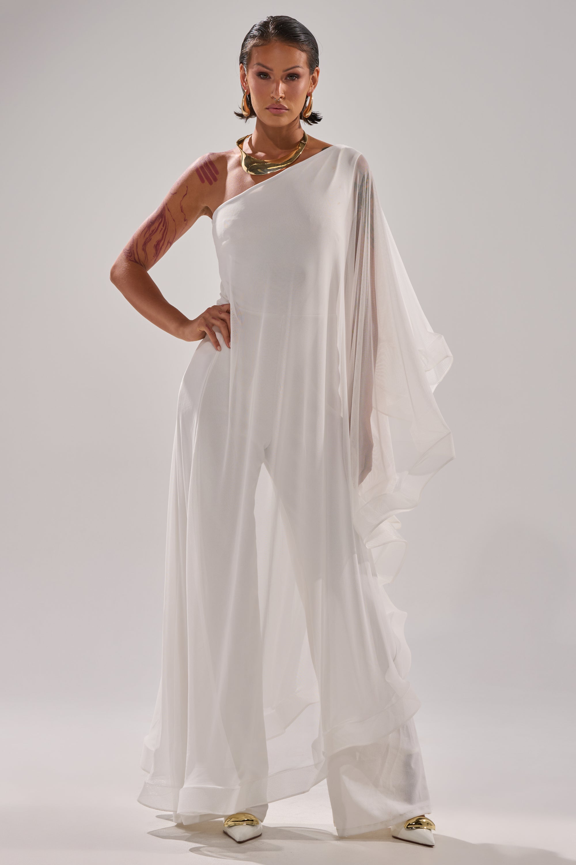 A person with short dark hair, visible arm tattoos, gold jewelry, and pointed gold shoes stands confidently in the SANDO MESH JUMPSUIT IN WHITE with wide legs and sheer mesh fabric against a plain light background. 2