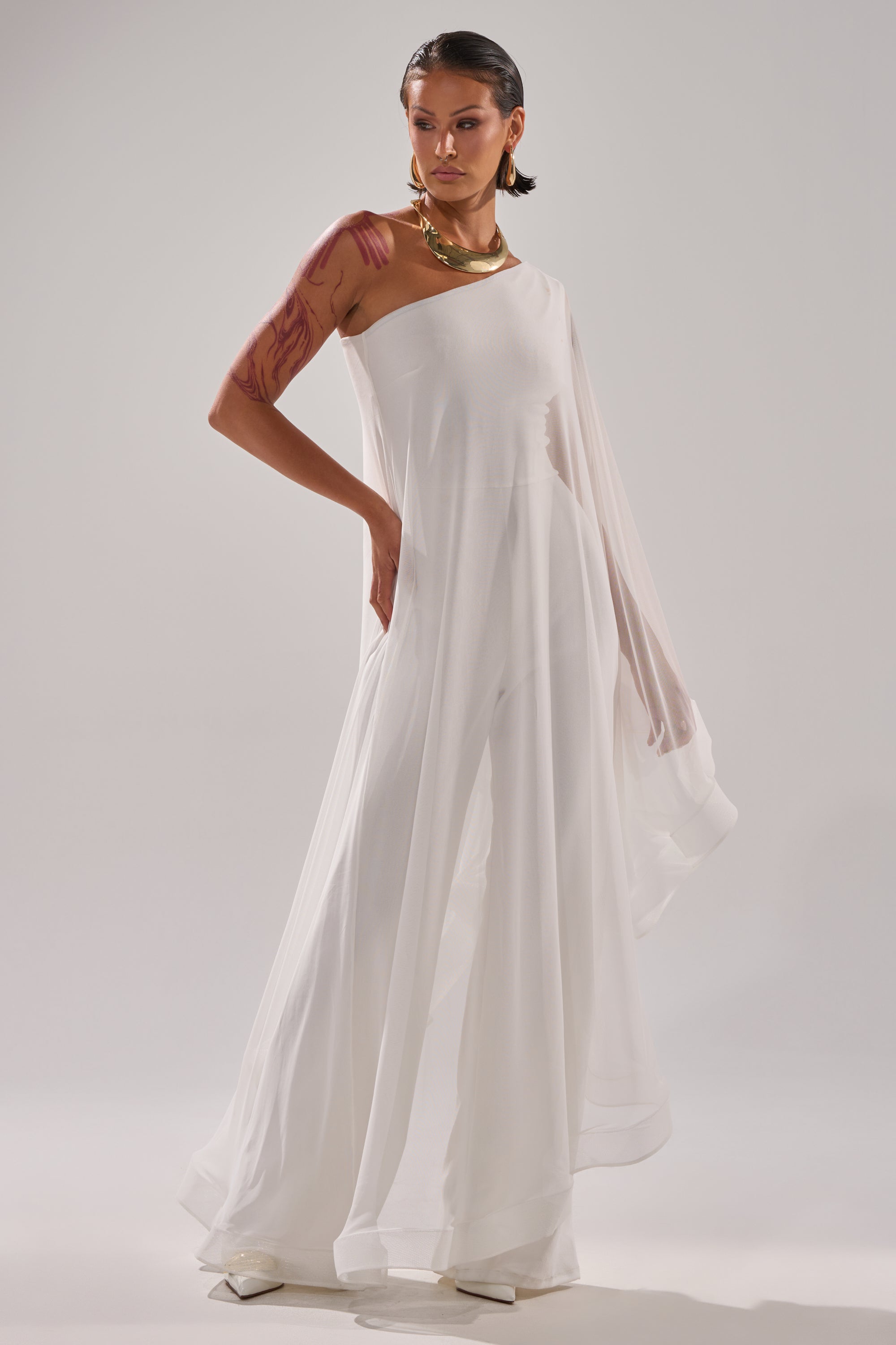 A person poses confidently in the SANDO MESH JUMPSUIT IN WHITE, featuring a flowing, one-shoulder design. They style it with slicked-back hair, bold gold jewelry, and visible body art on their right arm against a plain, light background. 3
