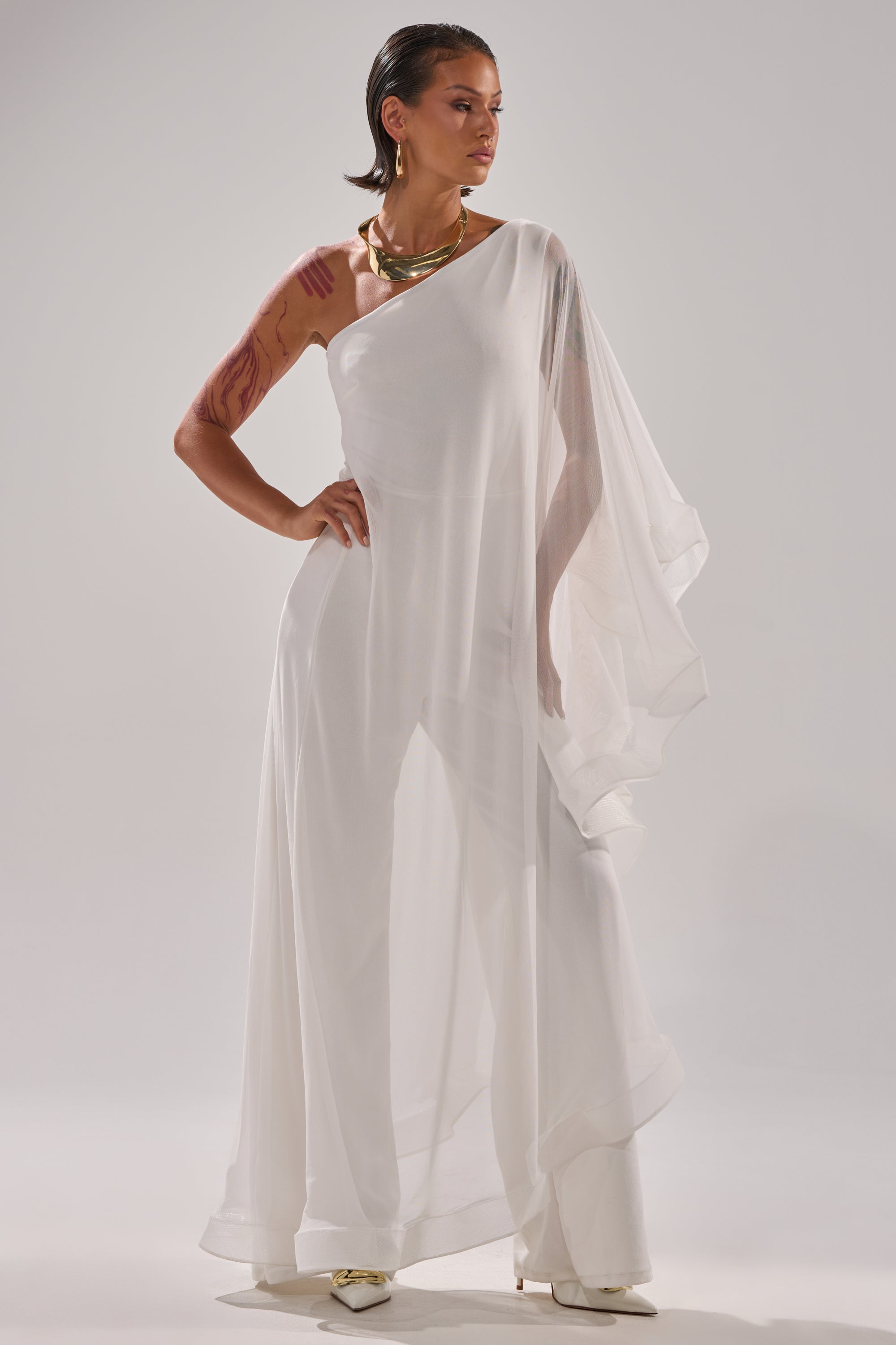 A woman stands confidently in the SANDO MESH JUMPSUIT IN WHITE, a sheer one-shoulder style with wide legs. She wears gold jewelry, has short dark hair and red markings on her arms, set against a plain light gray background. 4