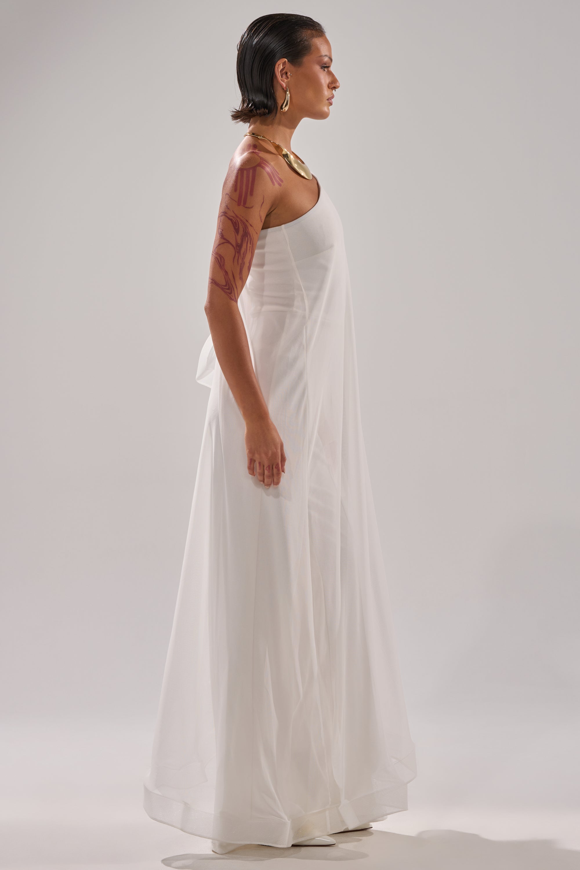 A person with slicked-back hair and a visible arm tattoo stands in profile, wearing the SANDO MESH JUMPSUIT IN WHITE and a gold necklace against a plain light background. 5