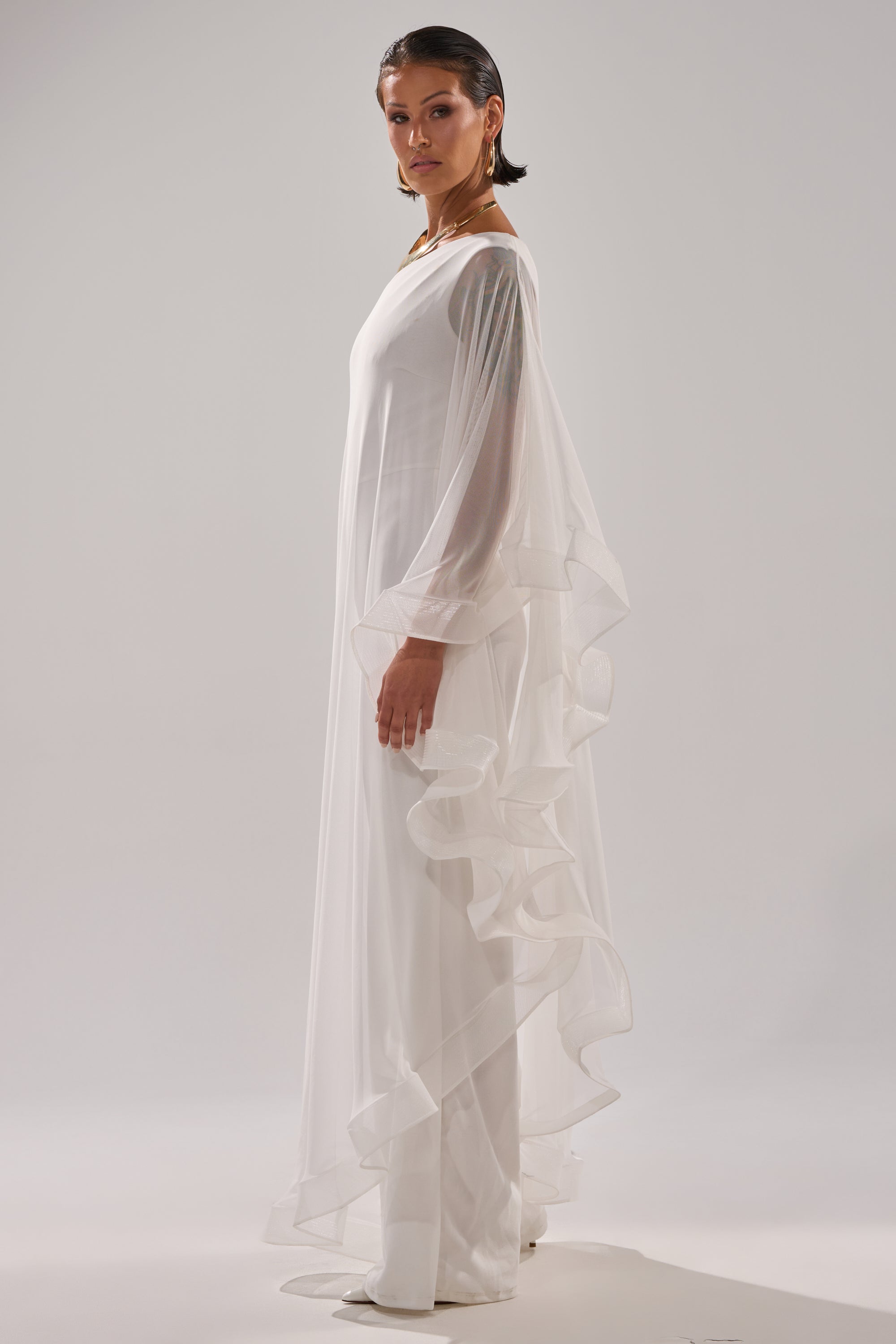 A woman stands sideways in the SANDO MESH JUMPSUIT IN WHITE, a sheer gown with ruffled edges layered over a chic white jumpsuit. Her hair is slicked back as she poses against a plain, light background. 6