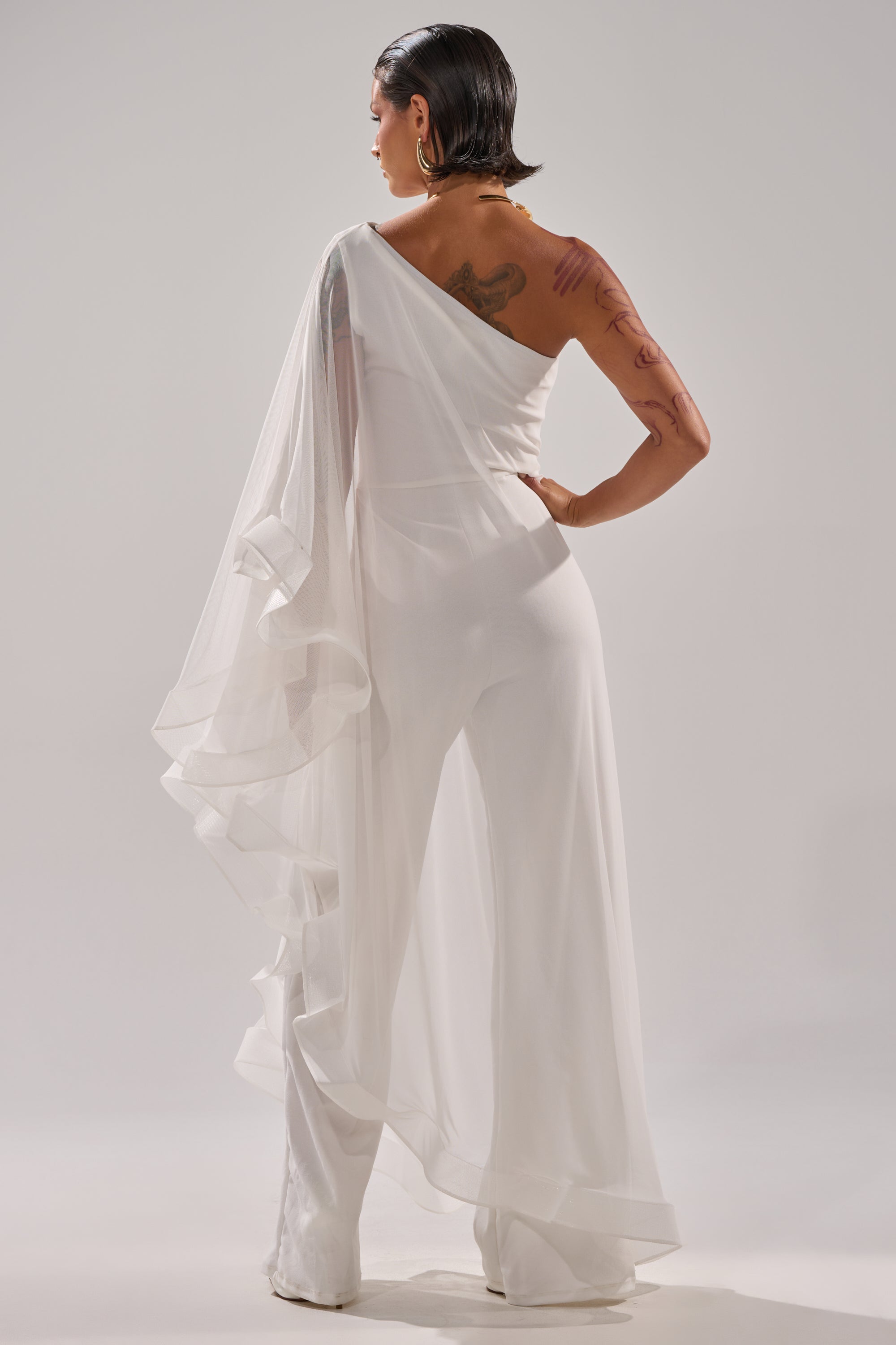 A person with short dark hair, tattoos on their upper back and arm, stands facing away in the SANDO MESH JUMPSUIT IN WHITE—an airy, one-shoulder design with sheer flowing fabric. The background is plain white. 7