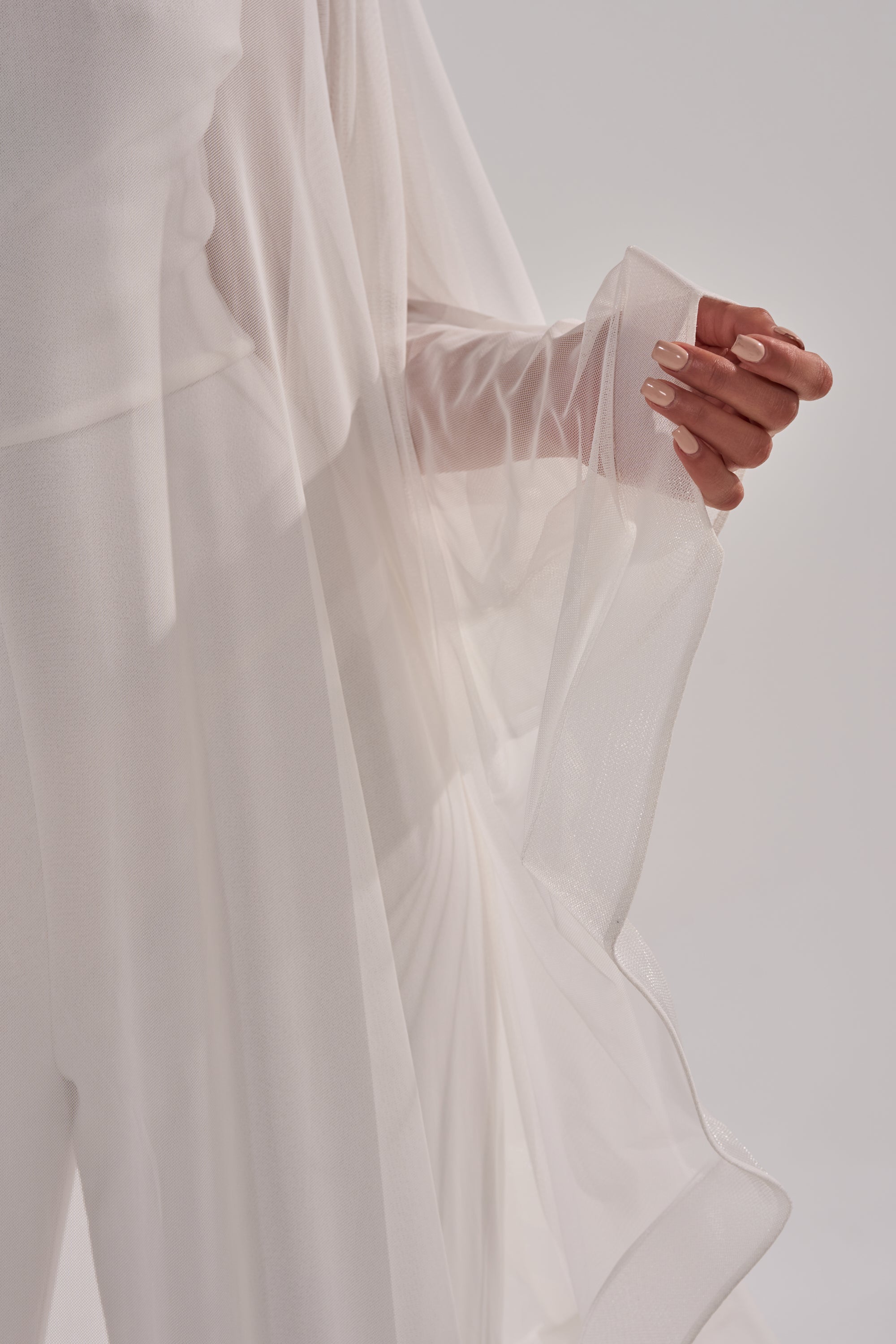 Wearing the SANDO MESH JUMPSUIT IN WHITE, a person gently holds the sheer fabric, revealing smooth, manicured nails against a softly lit, neutral background. 8