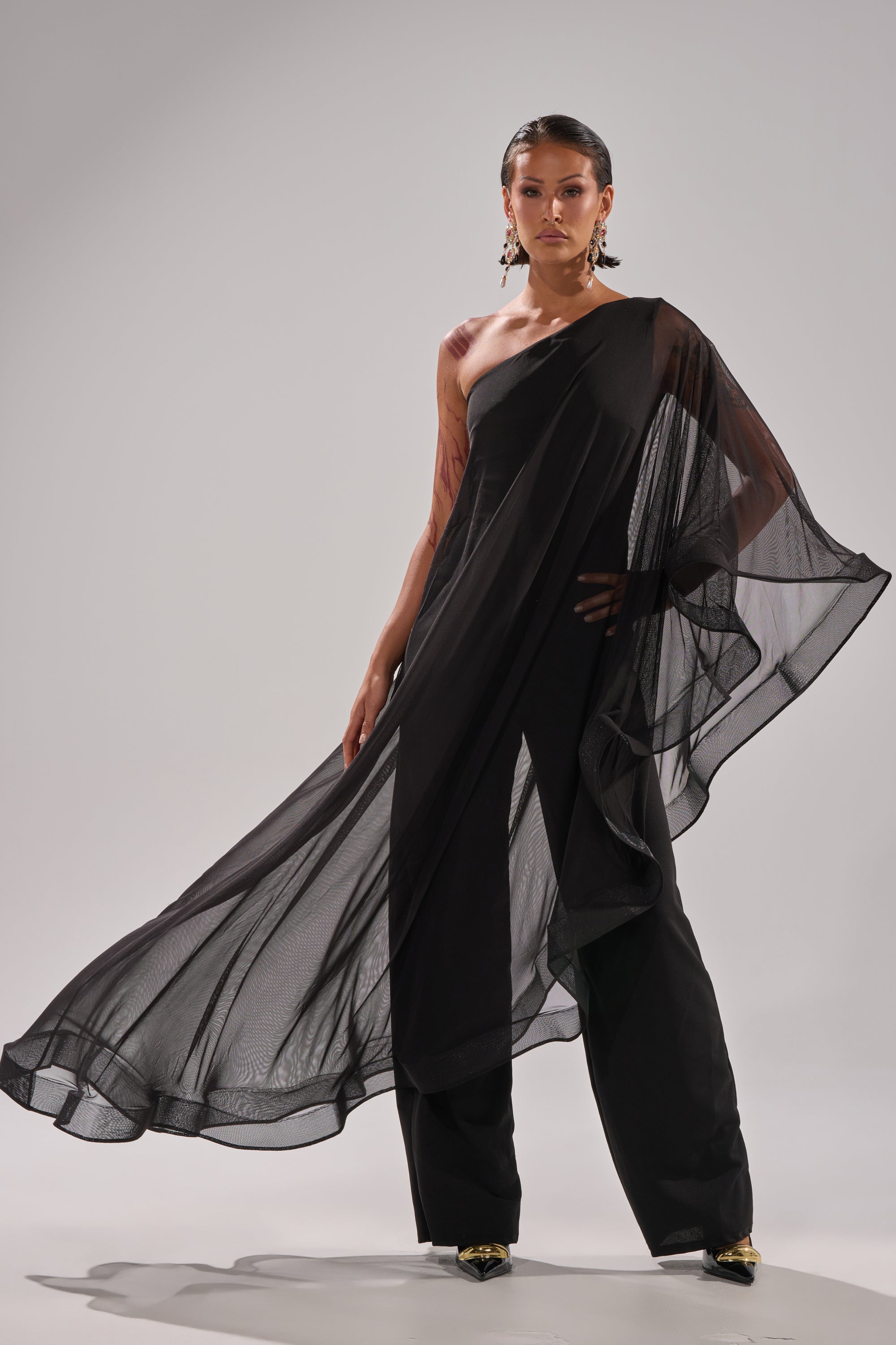 A person stands confidently in the SANDO MESH JUMPSUIT IN BLACK, featuring a chic one-shoulder design with a sheer mesh cape overlay and wide-leg pants, styled with statement earrings and pointed shoes against a plain light background.