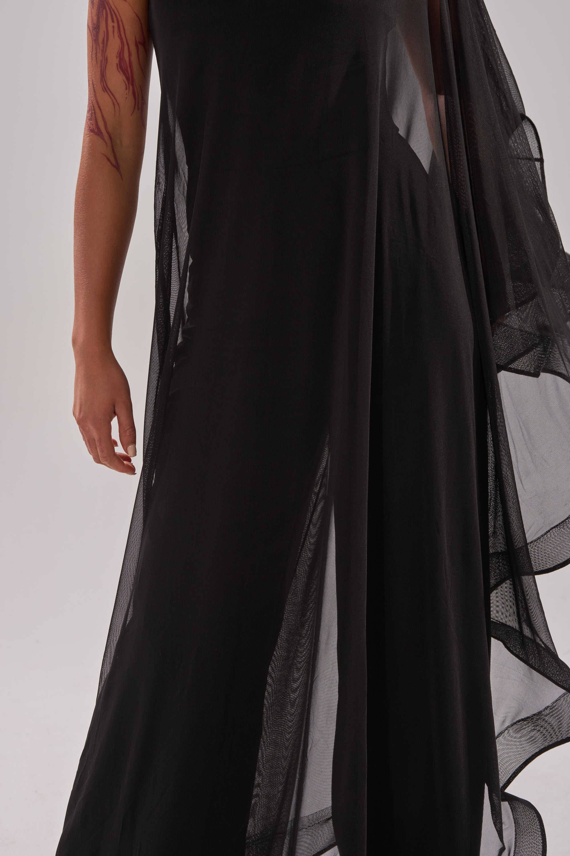 A person wears the SANDO MESH JUMPSUIT IN BLACK, a sheer flowing black outfit with their left arm exposed and a red tattoo visible, set against a plain light background.