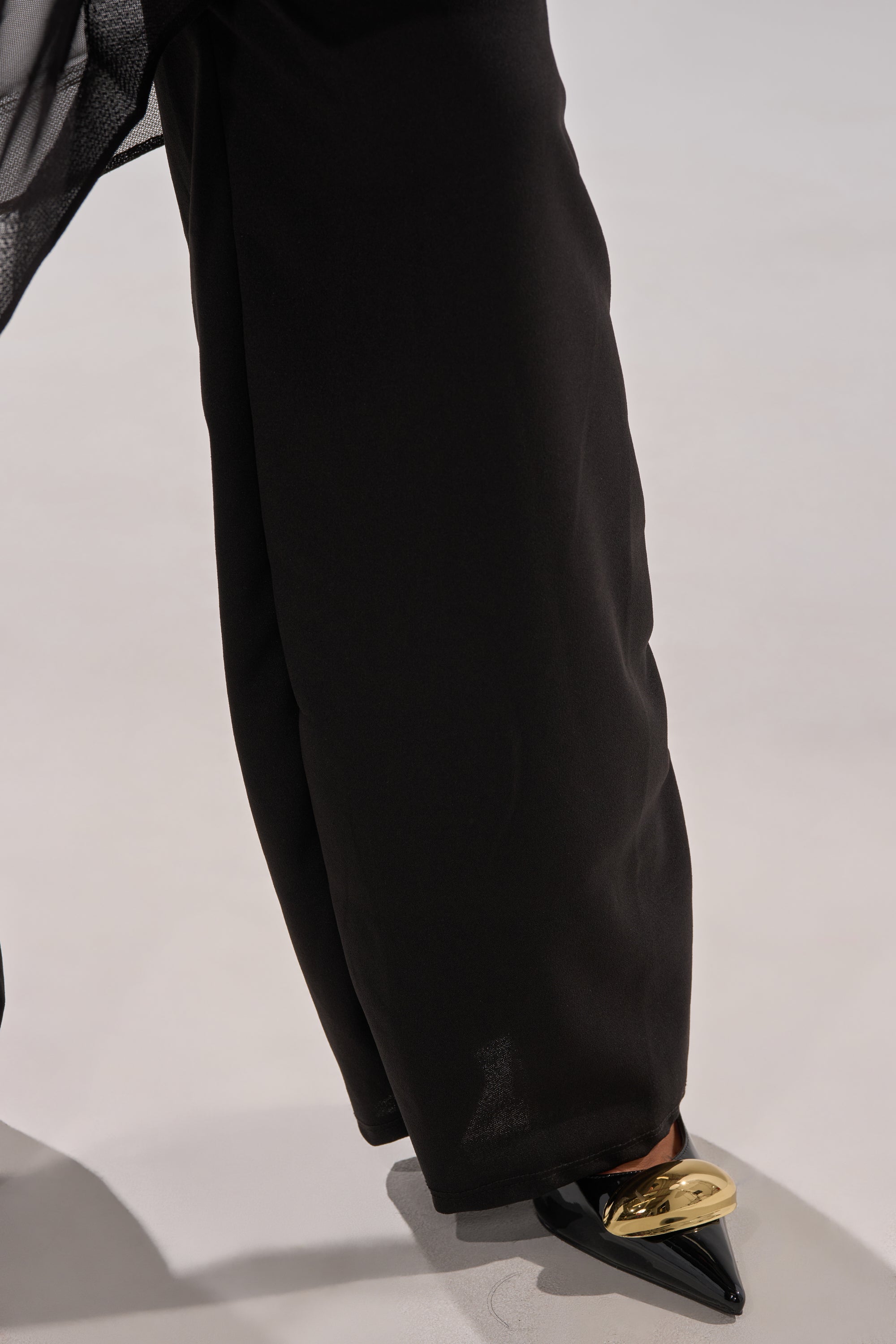 A person wears loose black pants and black pointed-toe shoes with a shiny gold accent, styled effortlessly with the SANDO MESH JUMPSUIT IN BLACK while standing on a light-colored surface.