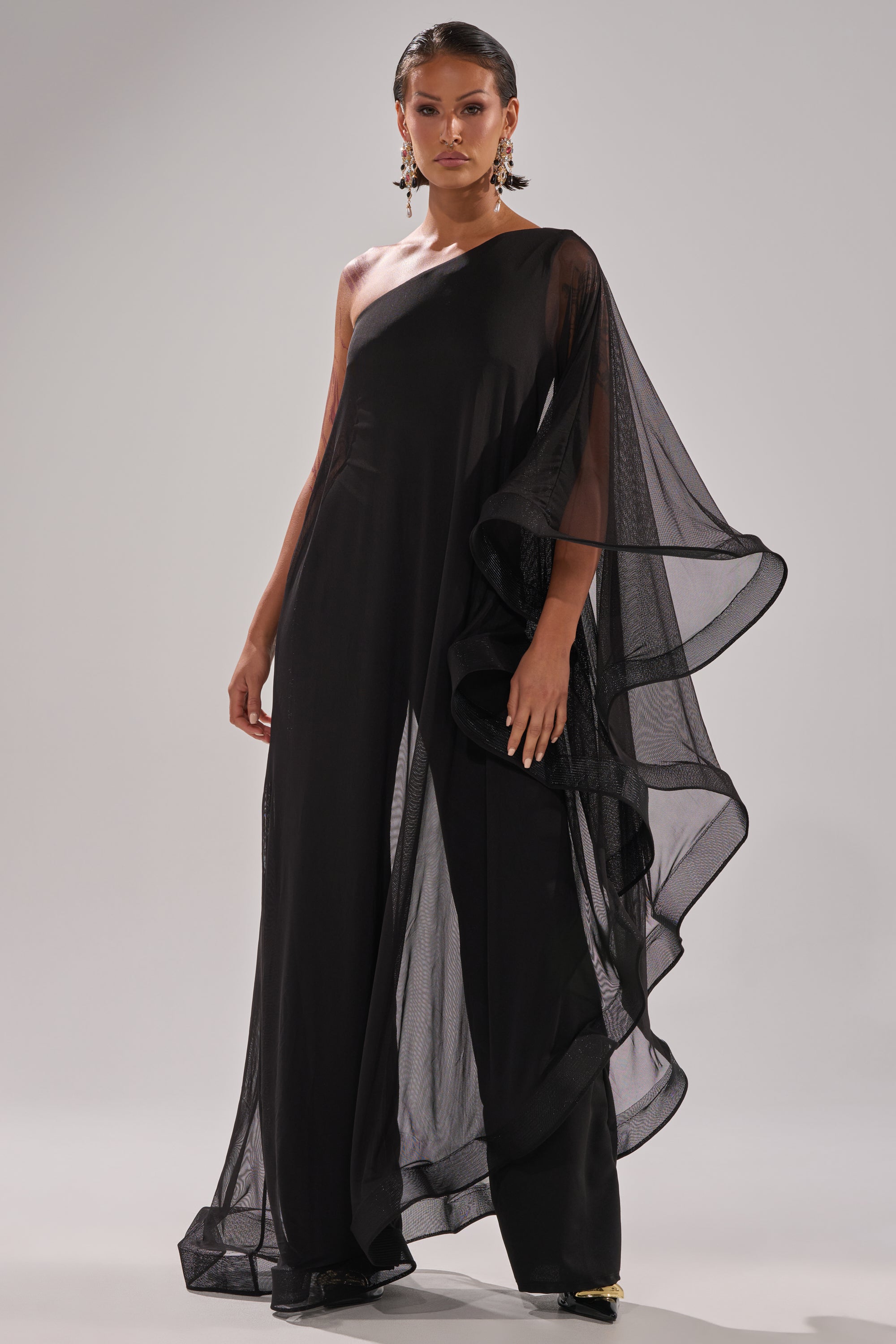 A woman stands confidently in the SANDO MESH JUMPSUIT IN BLACK, featuring sheer mesh fabric, ruffled details, and wide draping. She accessorizes with statement earrings and has slicked-back hair against a plain light-gray background.