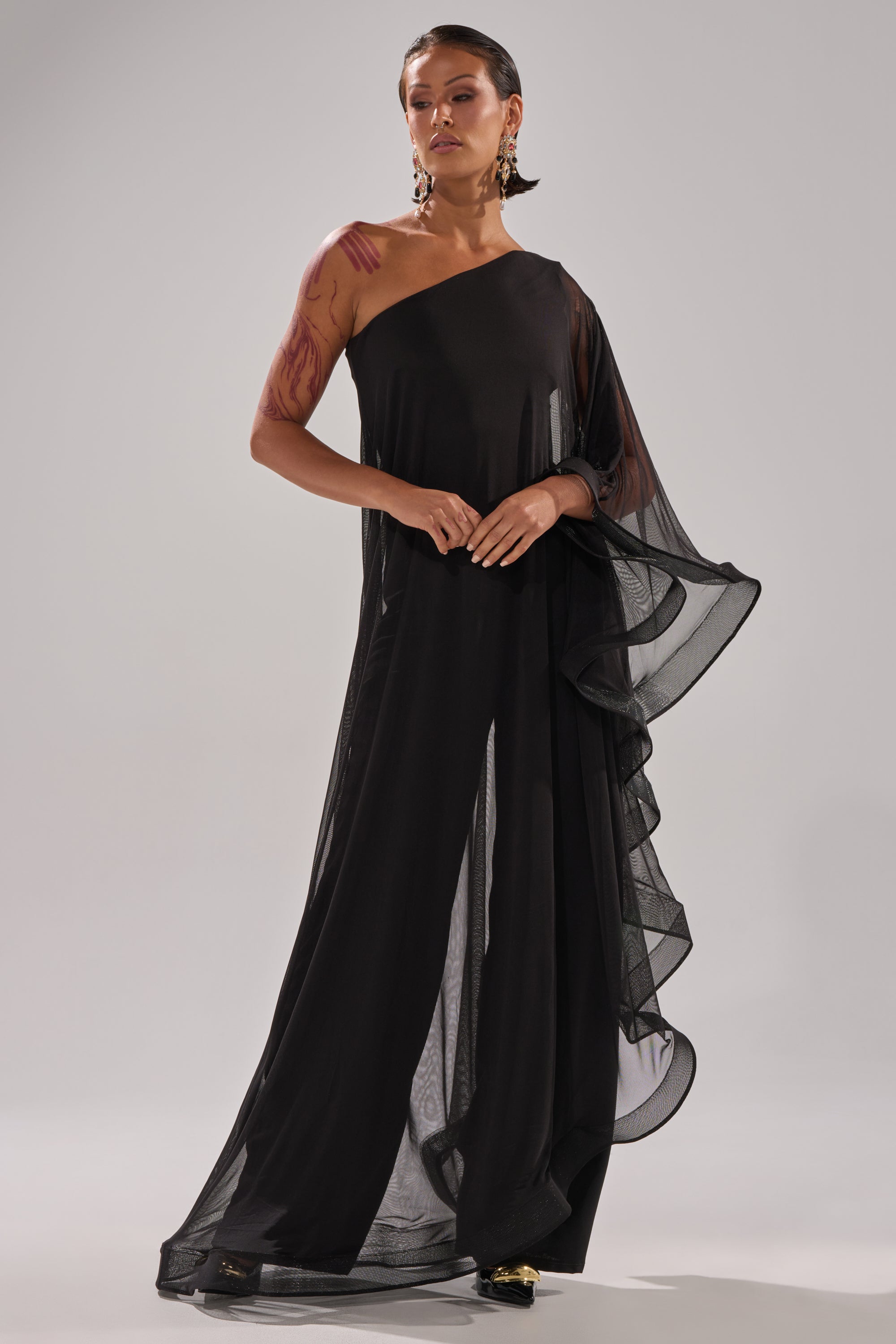 A person poses confidently in the SANDO MESH JUMPSUIT IN BLACK, featuring a flowing one-shoulder design with sheer mesh and ruffled draping. Statement earrings and visible tattoos complete their bold look against a plain background.