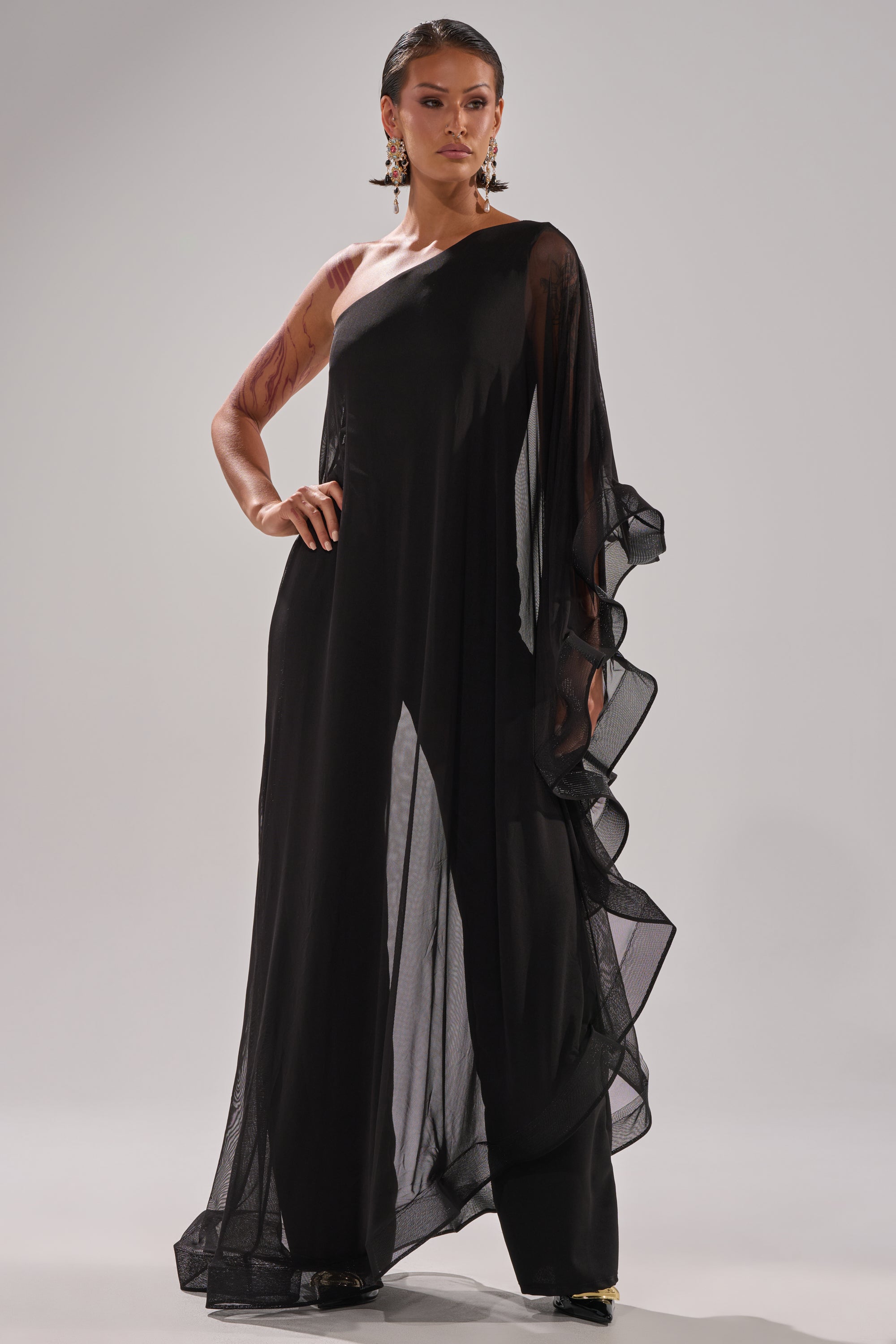 A woman poses confidently in the SANDO MESH JUMPSUIT IN BLACK, featuring a sheer one-shoulder design with a ruffled sleeve. She accessorizes with statement earrings and wears her hair slicked back, standing before a plain, light background.
